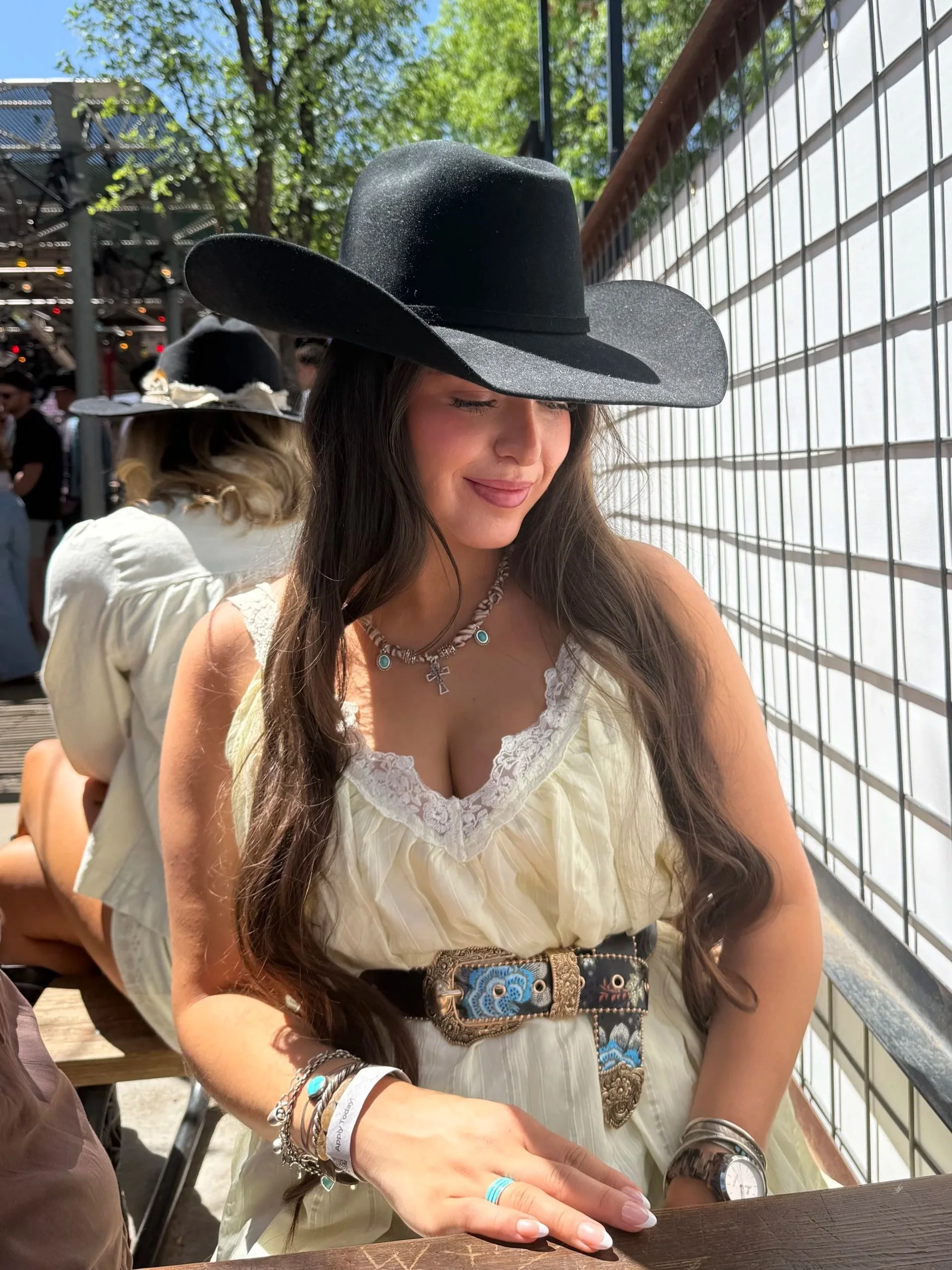 A woman with long dark hair wearing a black cowboy hat, a cream-colored dress with lace details, and bandana necklace, sitting outdoors near a wire fence, with trees and a clear sky in the background.