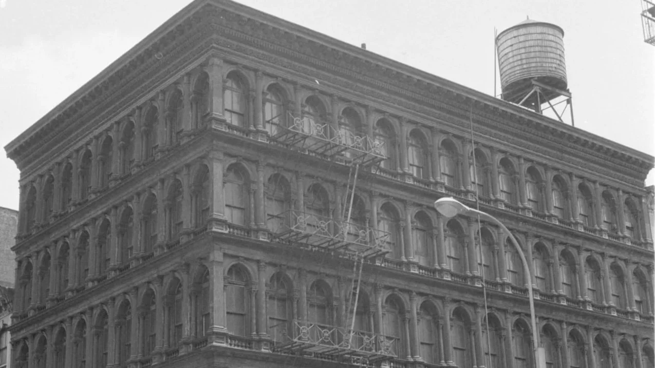 Black and white photo of a historic multi-story building with ornate architectural details, fire escapes on the side, a streetlight, and a water tower on the roof.