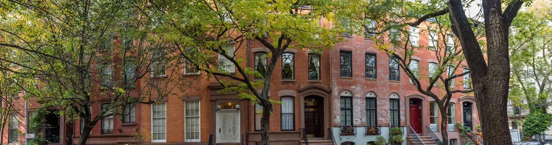 Street view of a row of brownstone apartment buildings with brick facades, front stoops, and trees with green leaves lining the sidewalk in an urban neighborhood.