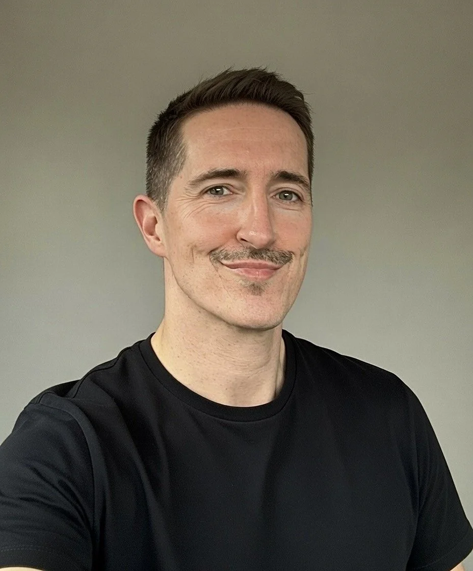 A smiling man with short brown hair, a mustache, and a goatee, wearing a black T-shirt, standing against a plain beige wall.
