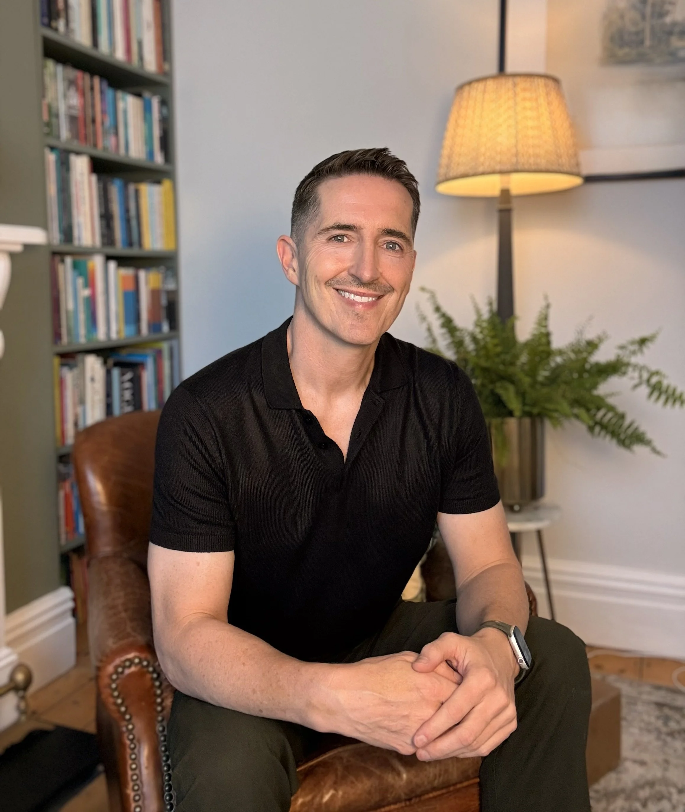 A smiling man with short brown hair, wearing a black shirt, seated on a brown leather armchair in a room with a bookshelf, a tall floor lamp, and houseplants behind him.