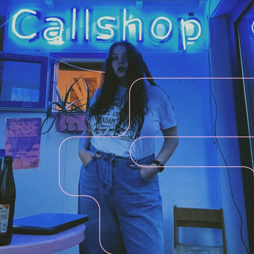 A woman with long dark curly hair wearing a graphic white t-shirt and high-waisted blue jeans standing inside a shop with a blue neon "Call shop" sign in the background, with her hands in her pockets, in a dimly lit room with blue lighting.