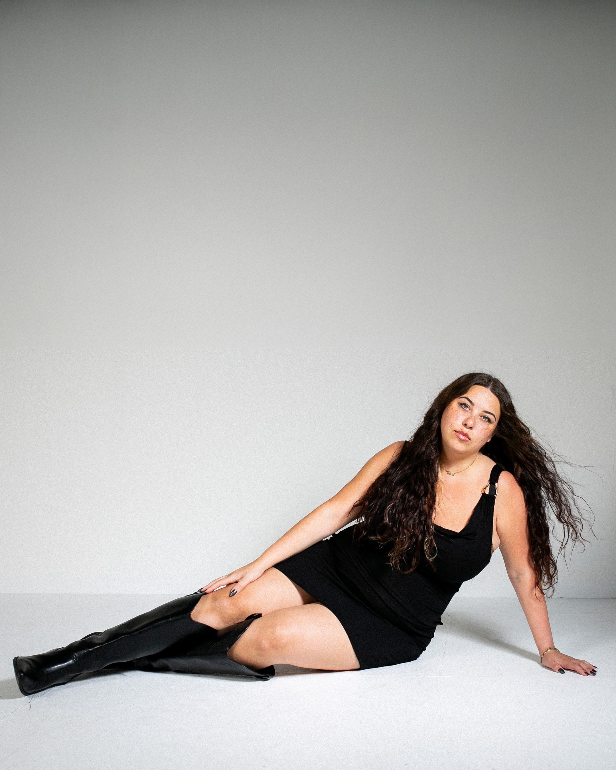 A woman with long wavy hair, wearing a black dress and black knee-high boots, sitting on the floor against a plain light gray backdrop.