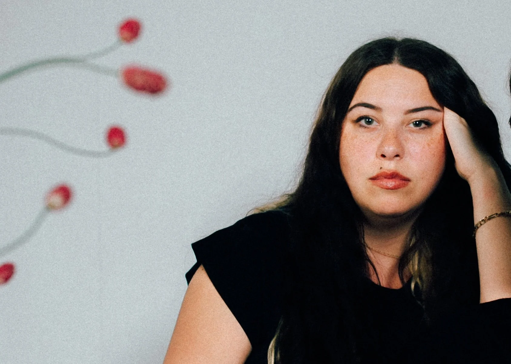 A woman with long dark hair and light skin, resting her head on her hand, sitting against a plain background with a blurred plant on the left.