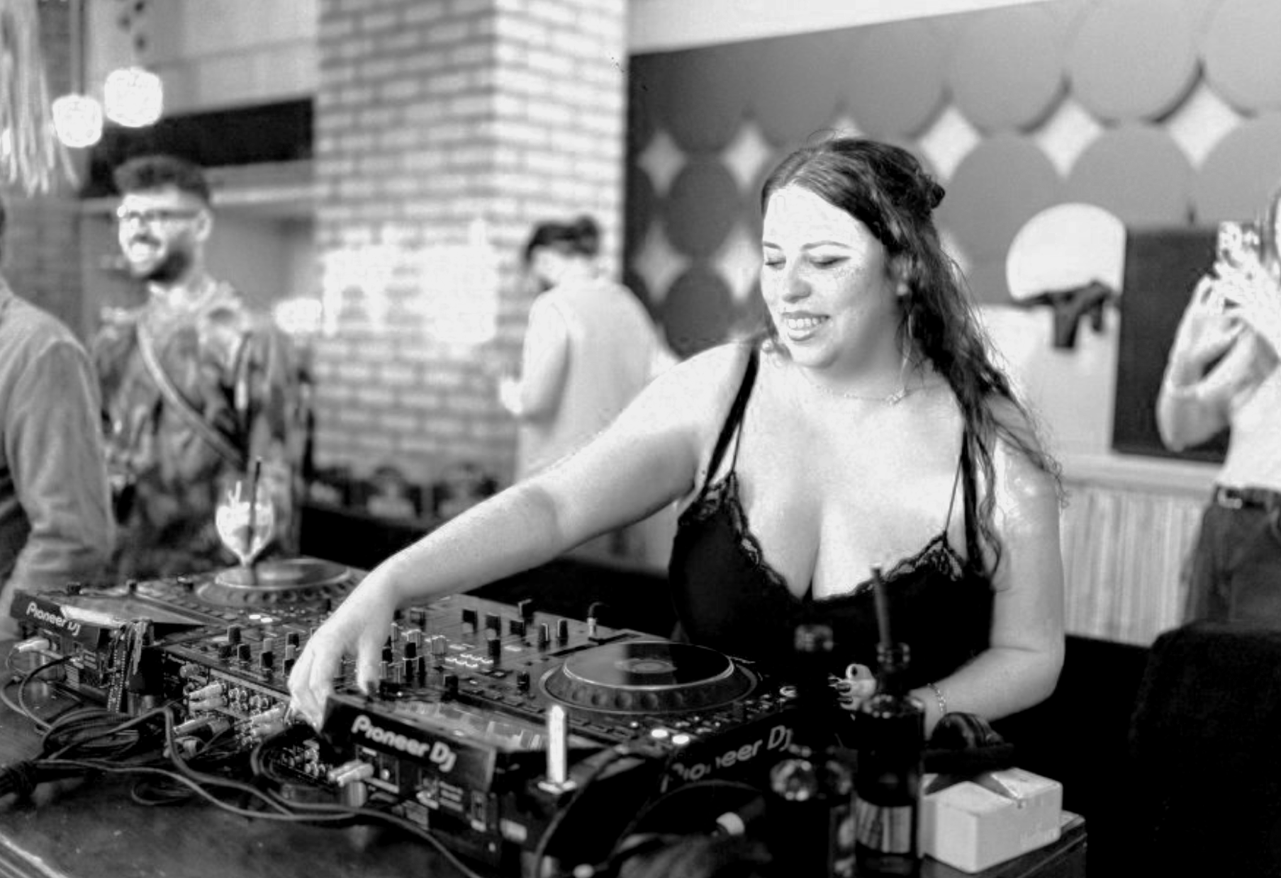 A woman DJ wearing a black dress with lace, smiling and mixing music on a DJ controller at a party, with people chatting in the background.