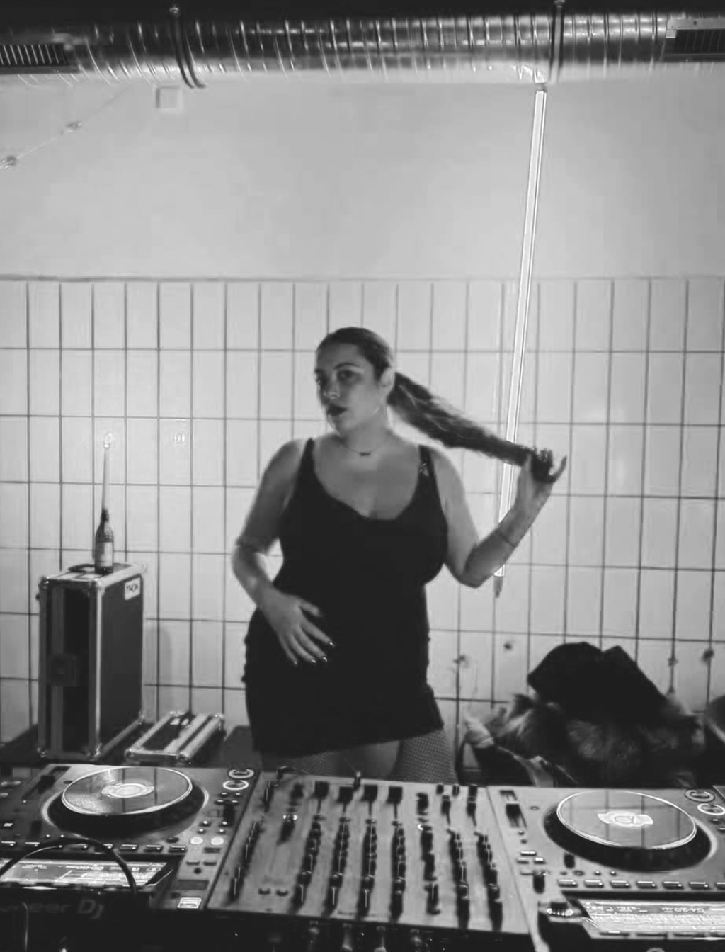 Black and white photo of a DJ woman in a black tank top and shorts posing with a long braid, standing behind DJ equipment in a tiled room with a vent and a bottle on a cabinet.
