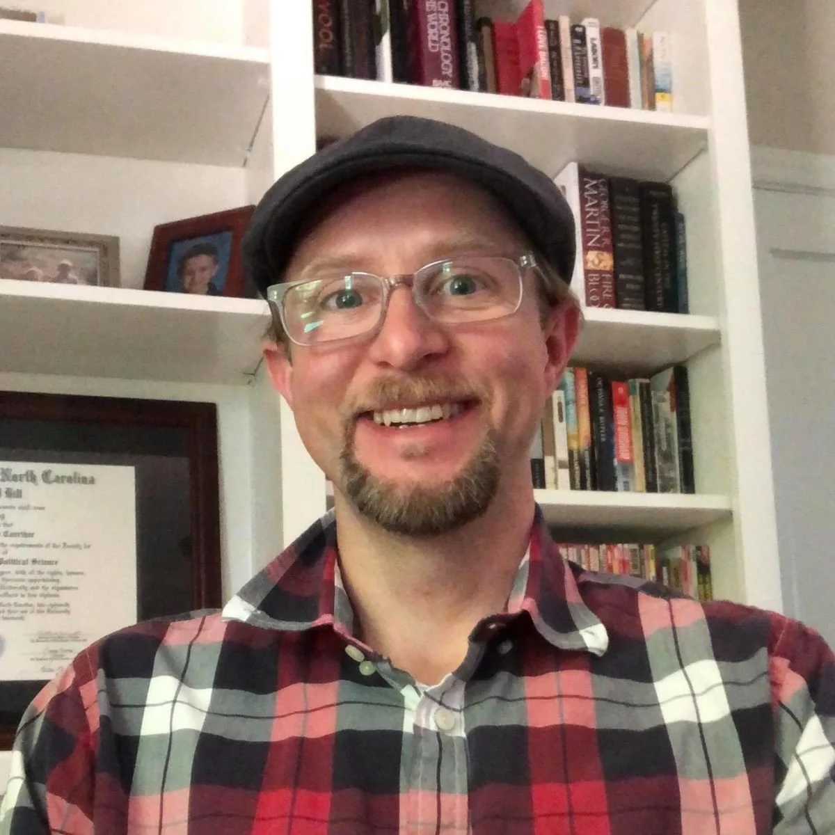 A man with glasses, a beard, and a mustache smiling in front of a bookshelf filled with books, wearing a cap and a red plaid shirt.