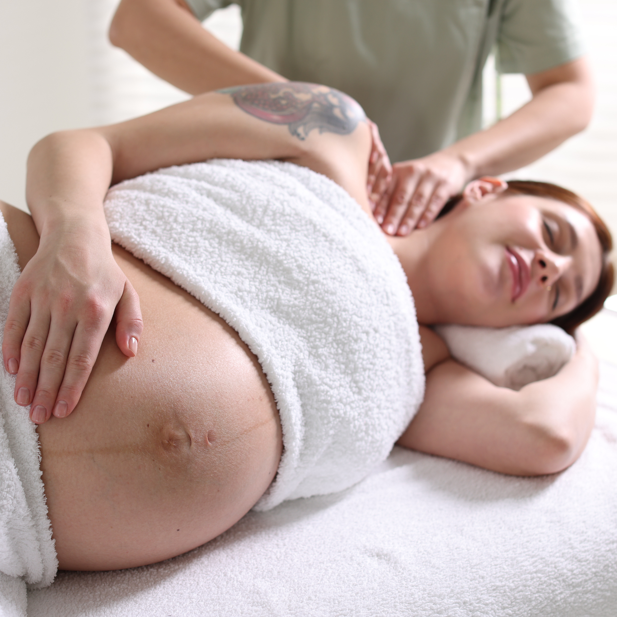 Person lying face down on a massage table receiving a massage, with a white towel covering their lower back and buttocks, and the therapist's hands on their back.