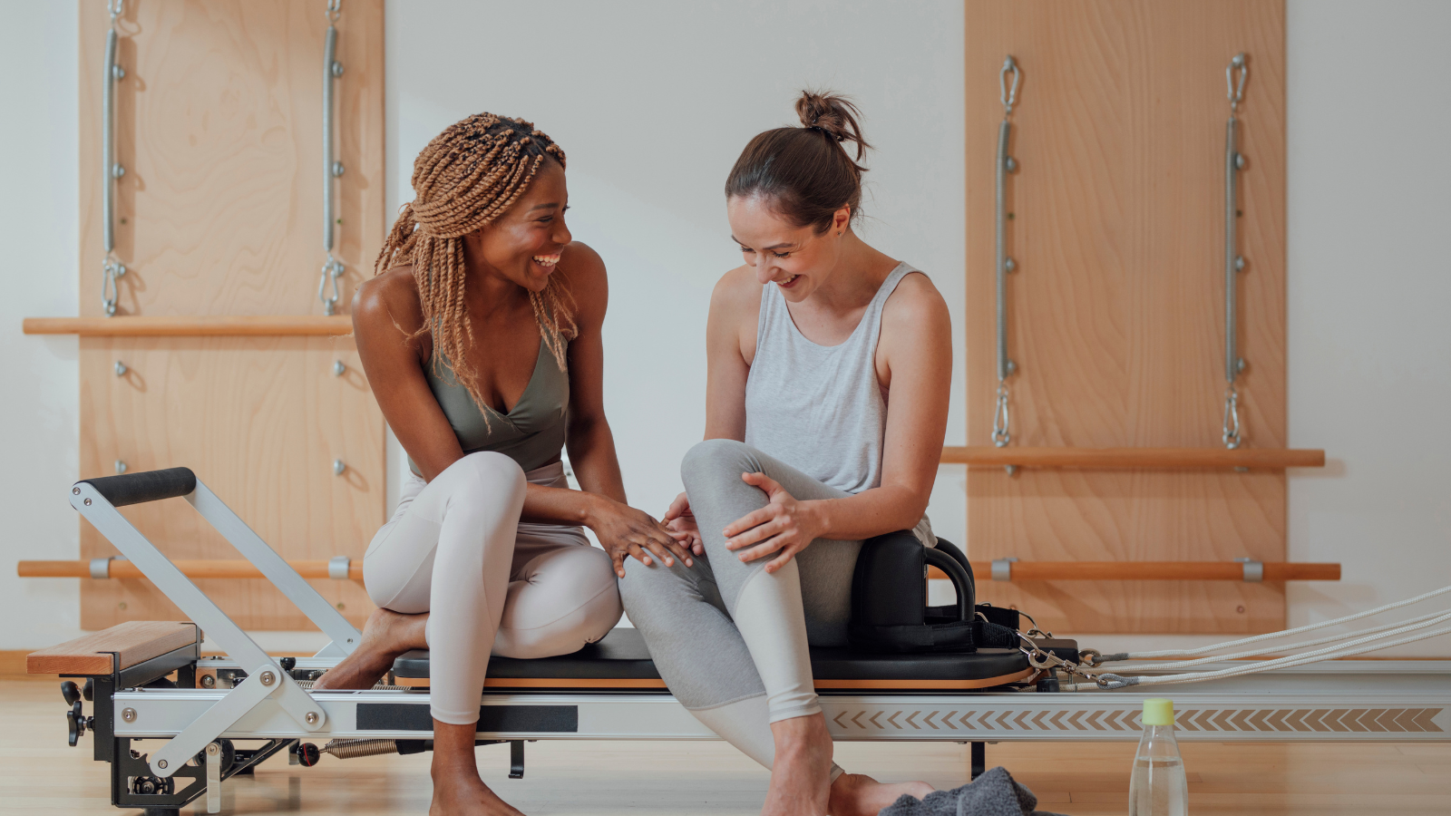Two women laughing and supporting each other during a pilates session in a studio with wooden wall bars in the background.