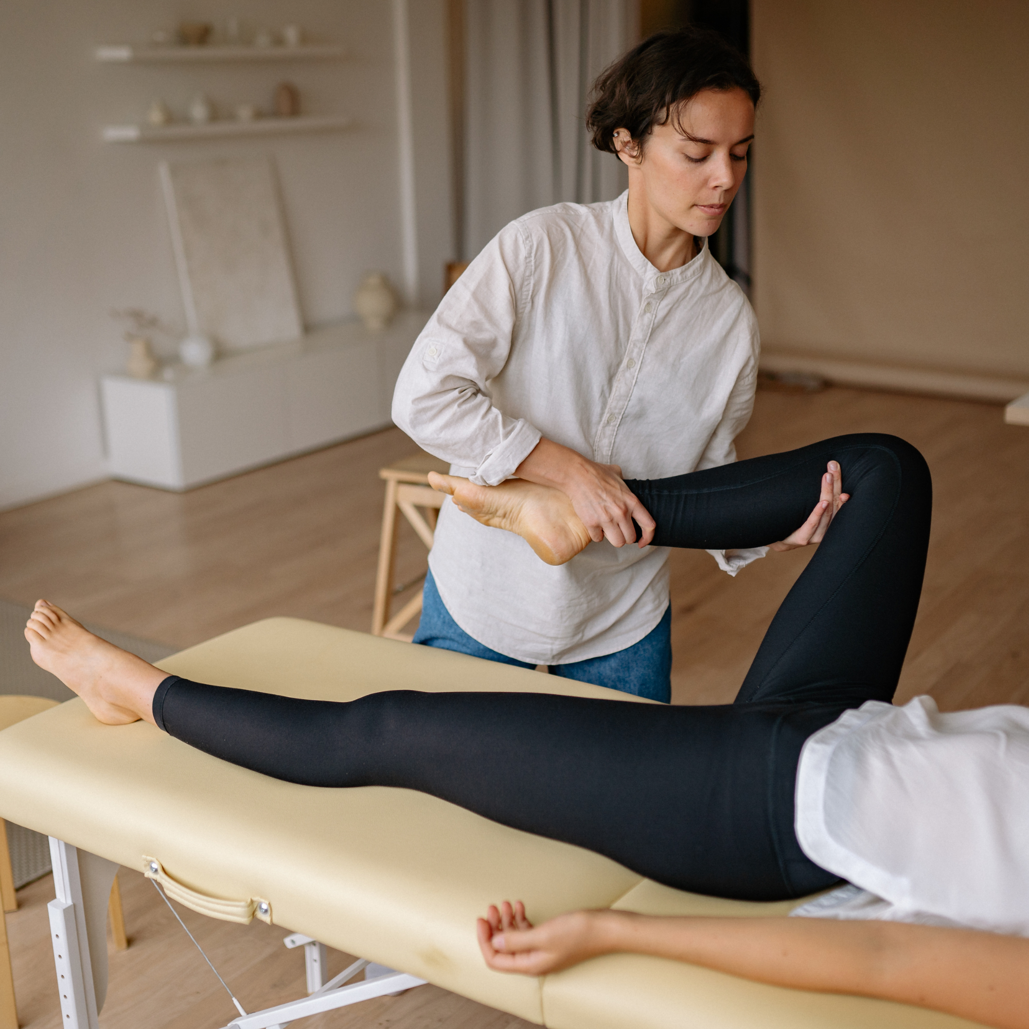 A woman in a white shirt is performing physical therapy on another person's leg while sitting in a room with a beige and brown color scheme. The room has an art easel, shelves with decorative items, and neutral curtains.