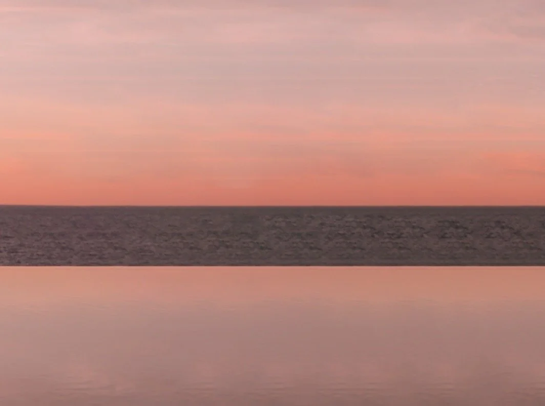 Calm body of water reflecting a pink and orange sunset sky with a dark horizon line.