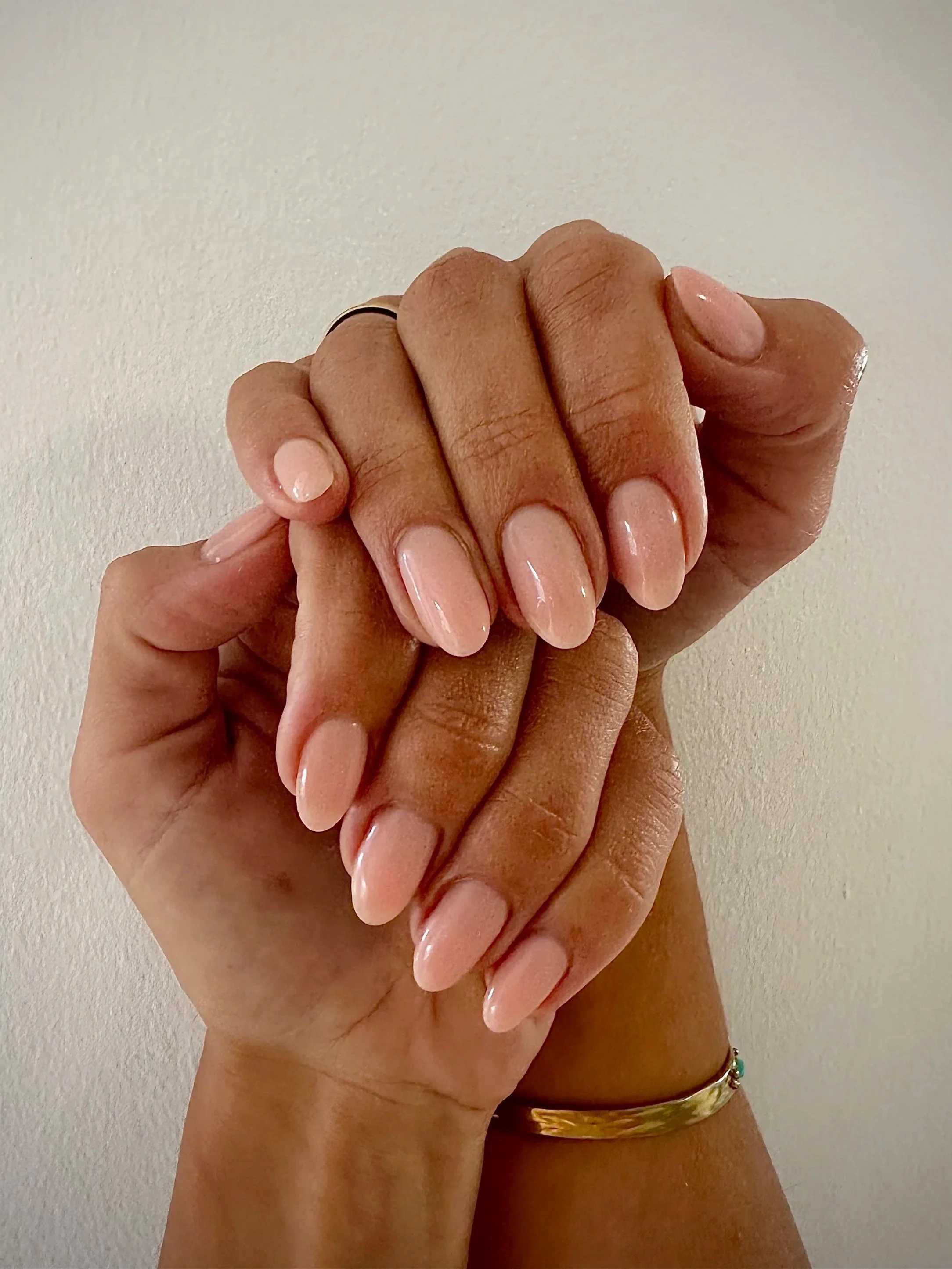 Two hands with manicured nails clasped together, one hand wearing a gold bracelet and a black ring, the nails are painted with a light pink nail polish.