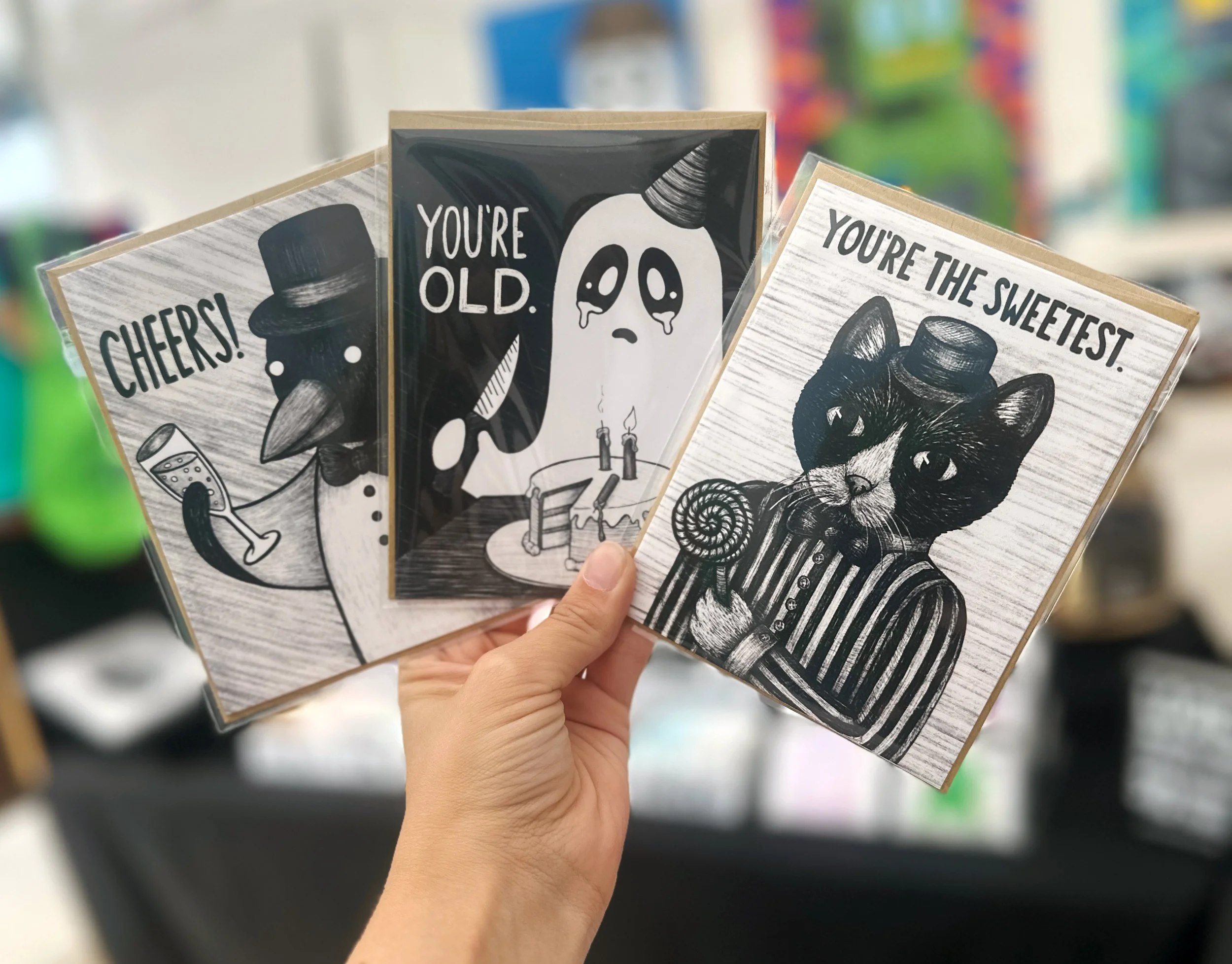 Image of greeting cards. One has a penguin raising a glass with the phrase "Cheers!" The second is of a crying ghost cutting into a birthday cake with the phrase "You're Old." And the third is of a cat with a lollipop saying "You're The Sweetest."