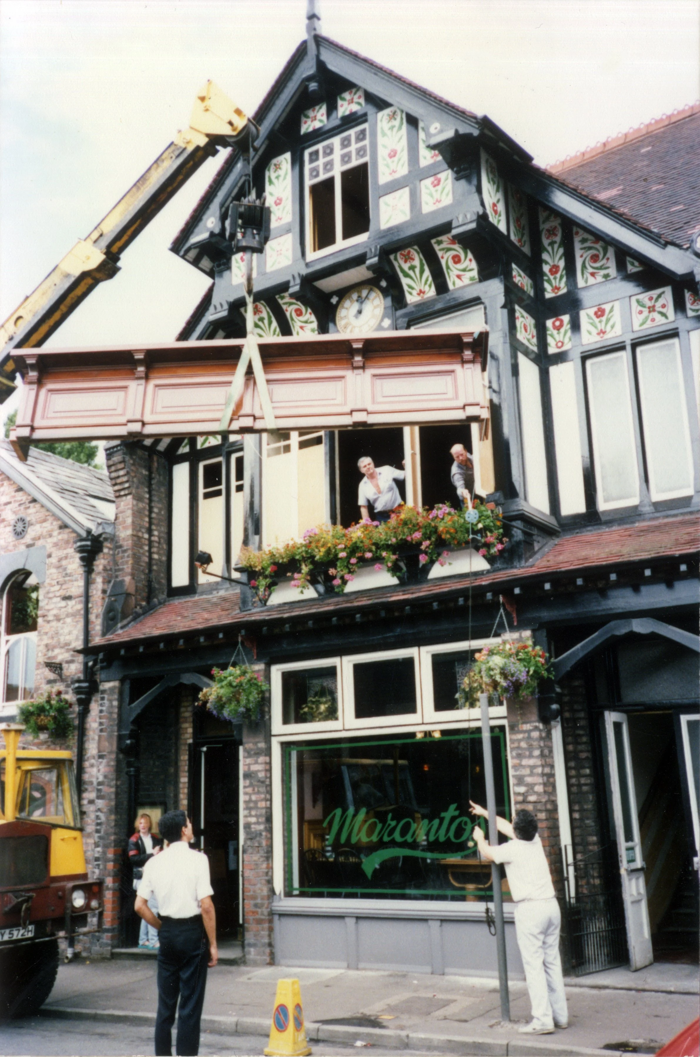 A building with traditional European architecture, featuring decorative painted woodwork and hanging flower baskets. A crane is lifting a rectangular balcony or sign frame above the entrance, with people standing nearby. The scene appears to be in a busy urban area.