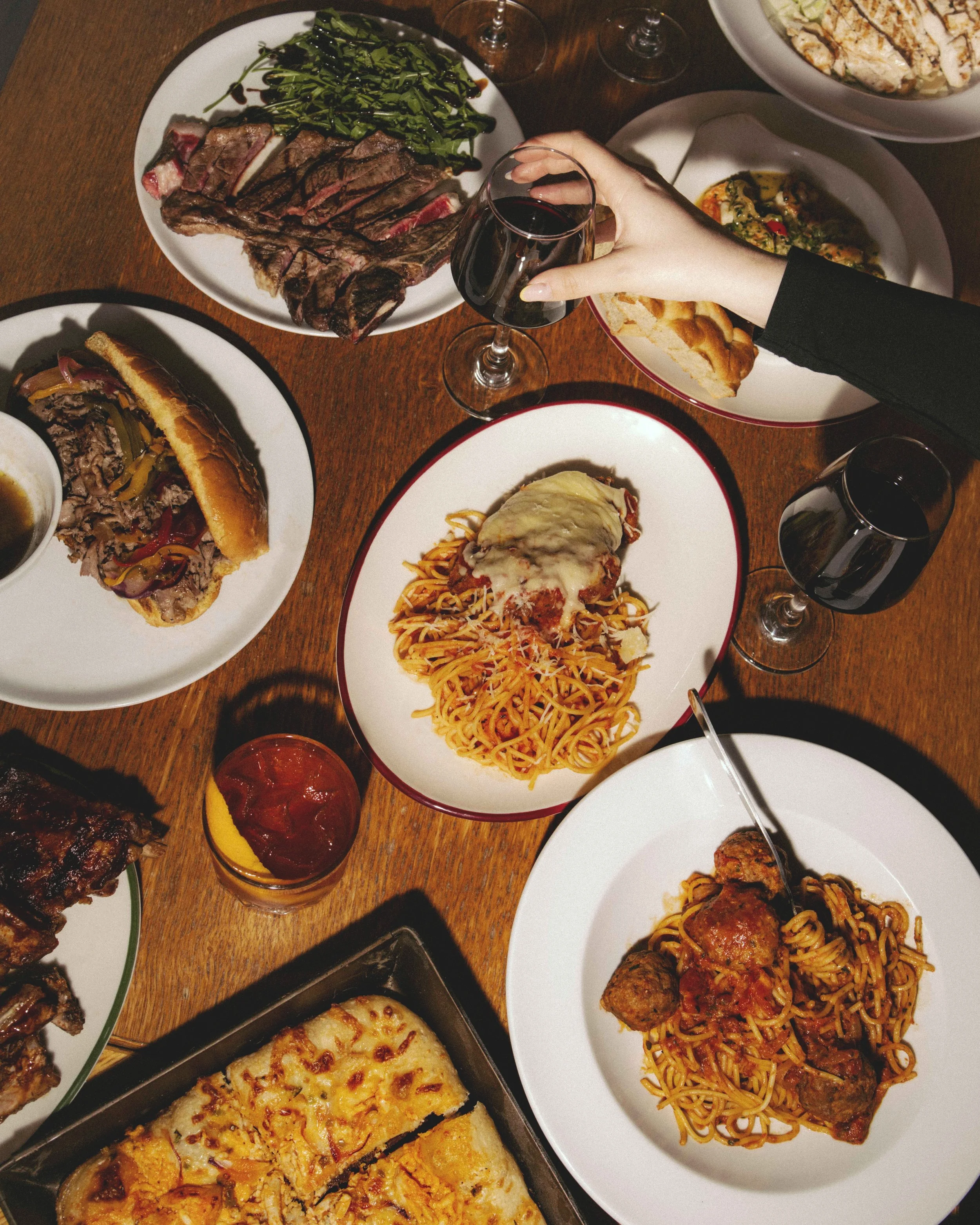 A dinner table with various dishes, including sliced steak, a beef sandwich, spaghetti with meatballs, pizza, and salad, along with glasses of red wine.
