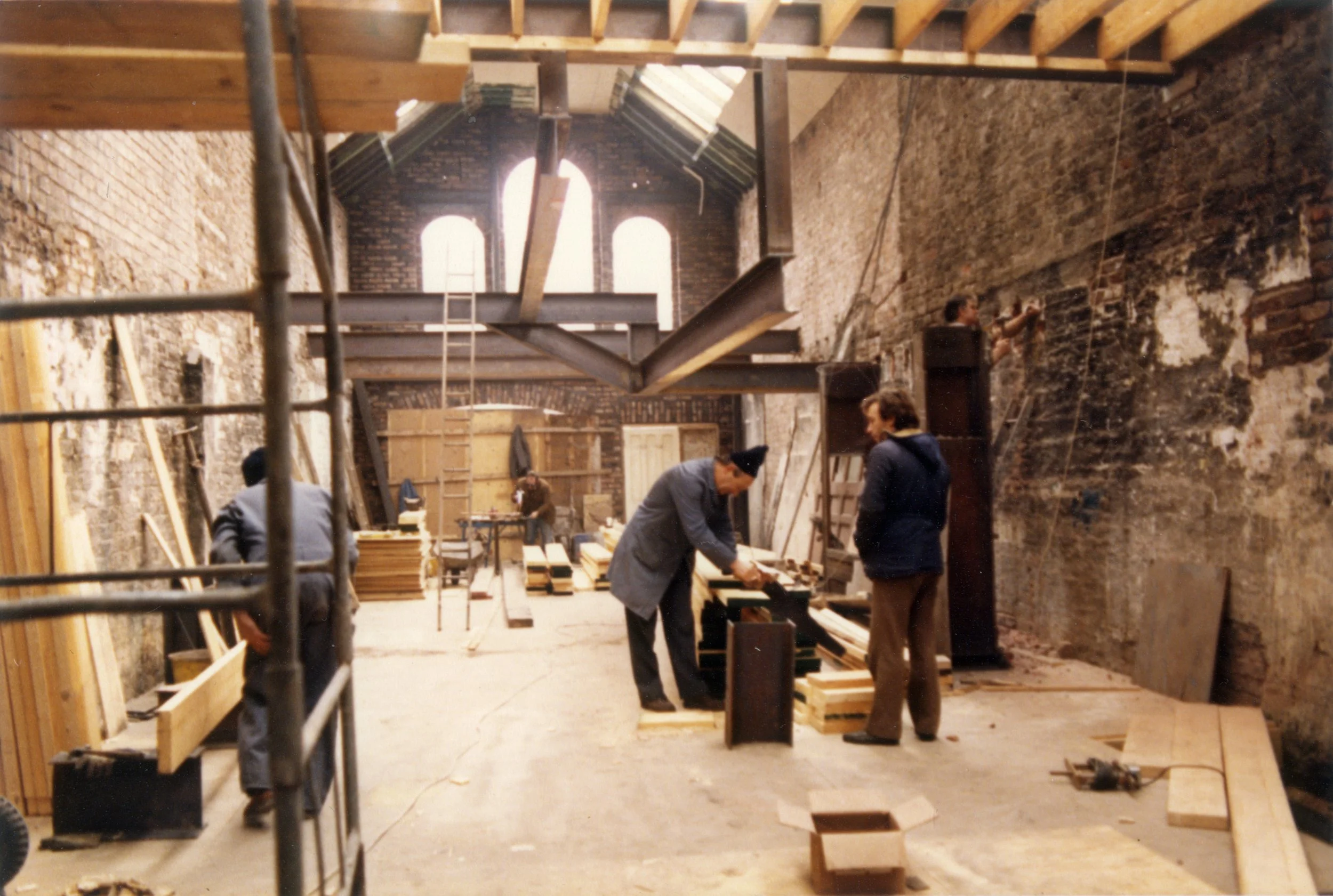 Interior of a building under renovation, with exposed brick walls, wooden beams, and multiple workers constructing or renovating the space, including scaffolding and construction materials.