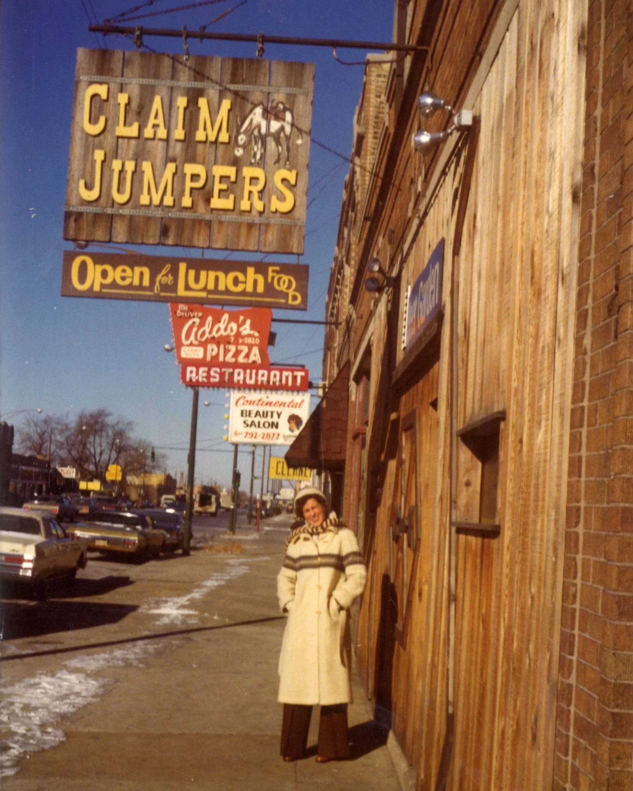 A woman stands on a sidewalk in front of a brick building with ice patches on the ground, wearing a cream-colored coat and a matching hat. There are signs above and beside her, including a large wooden sign for 'Claim Jumpers' with an illustration of a person and horse, and others advertising a pizza restaurant and a beauty salon. The street has parked cars, and the sky is clear with some trees in the background.