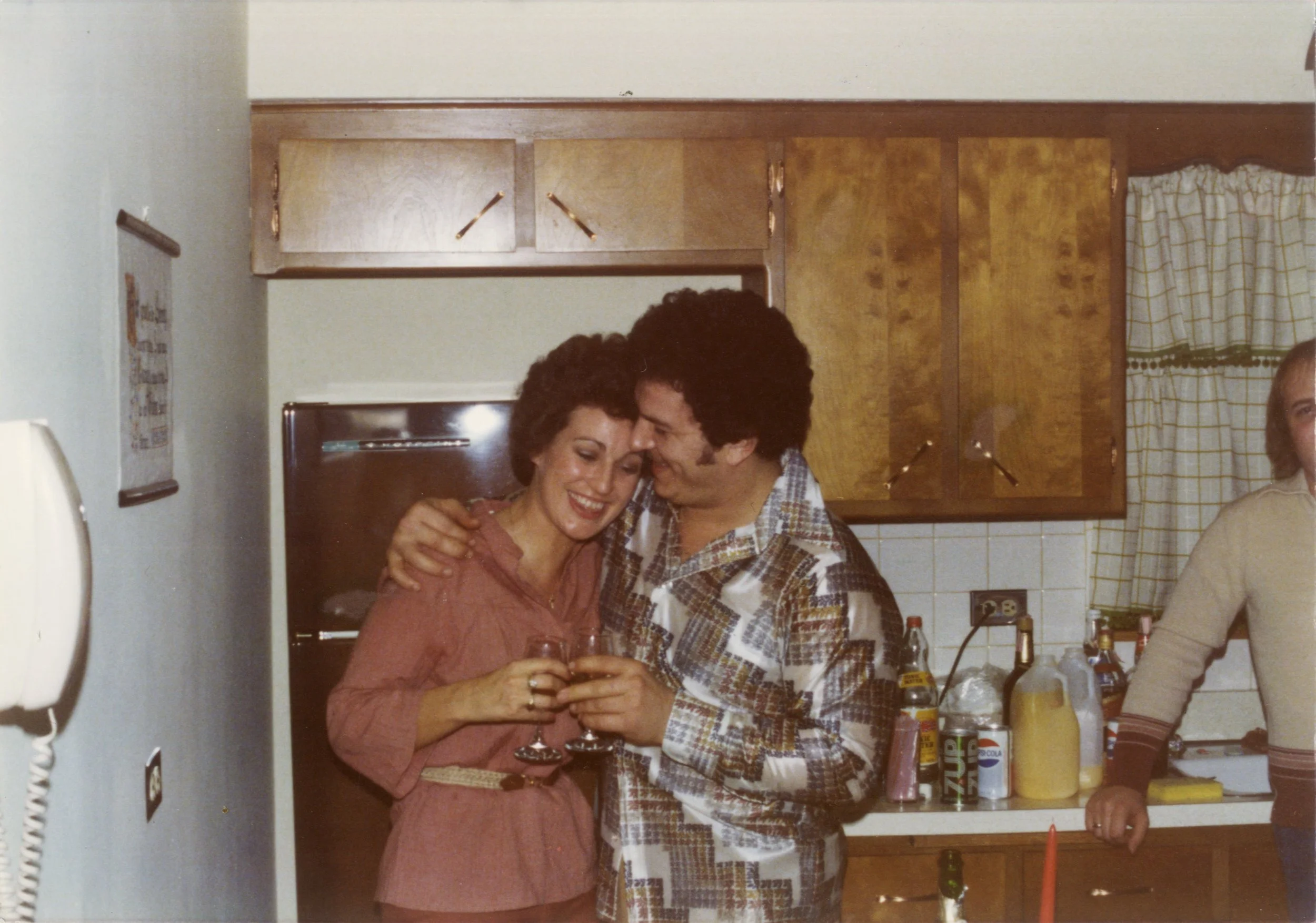 A woman in a pink dress and a man in plaid clothing are smiling and embracing, holding glasses of wine in a kitchen with wooden cabinets, curtains, and various condiments on the counter.