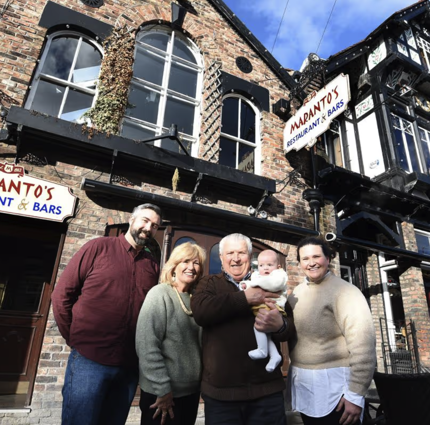 Group of five adults and one baby standing outside a brick restaurant called Manto's with signs visible, smiling at the camera with a blue sky overhead.