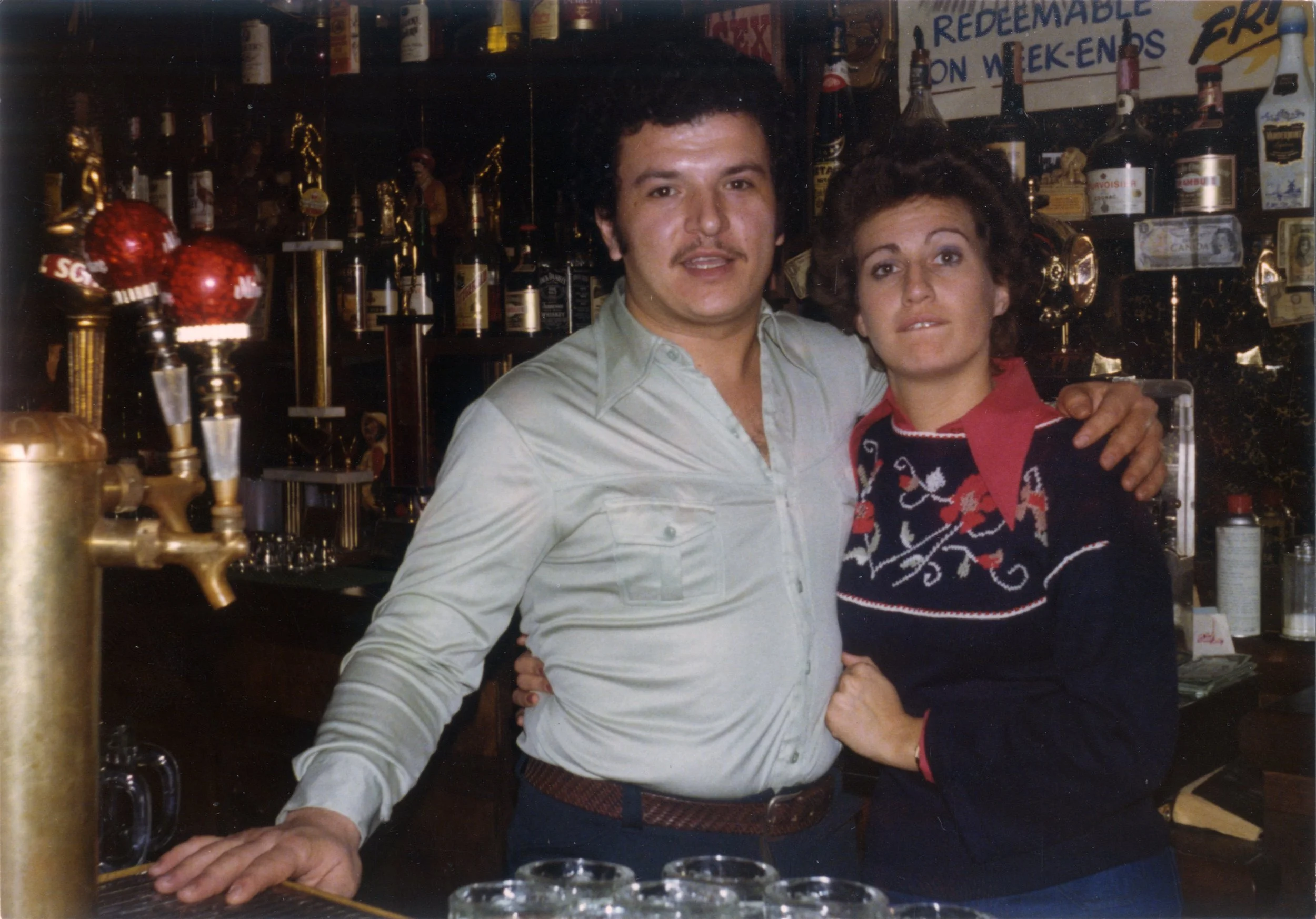 A man and a woman standing together in a bar, with the man wearing a light-colored shirt and the woman wearing a Christmas sweater, surrounded by bottles of alcohol on shelves in the background.