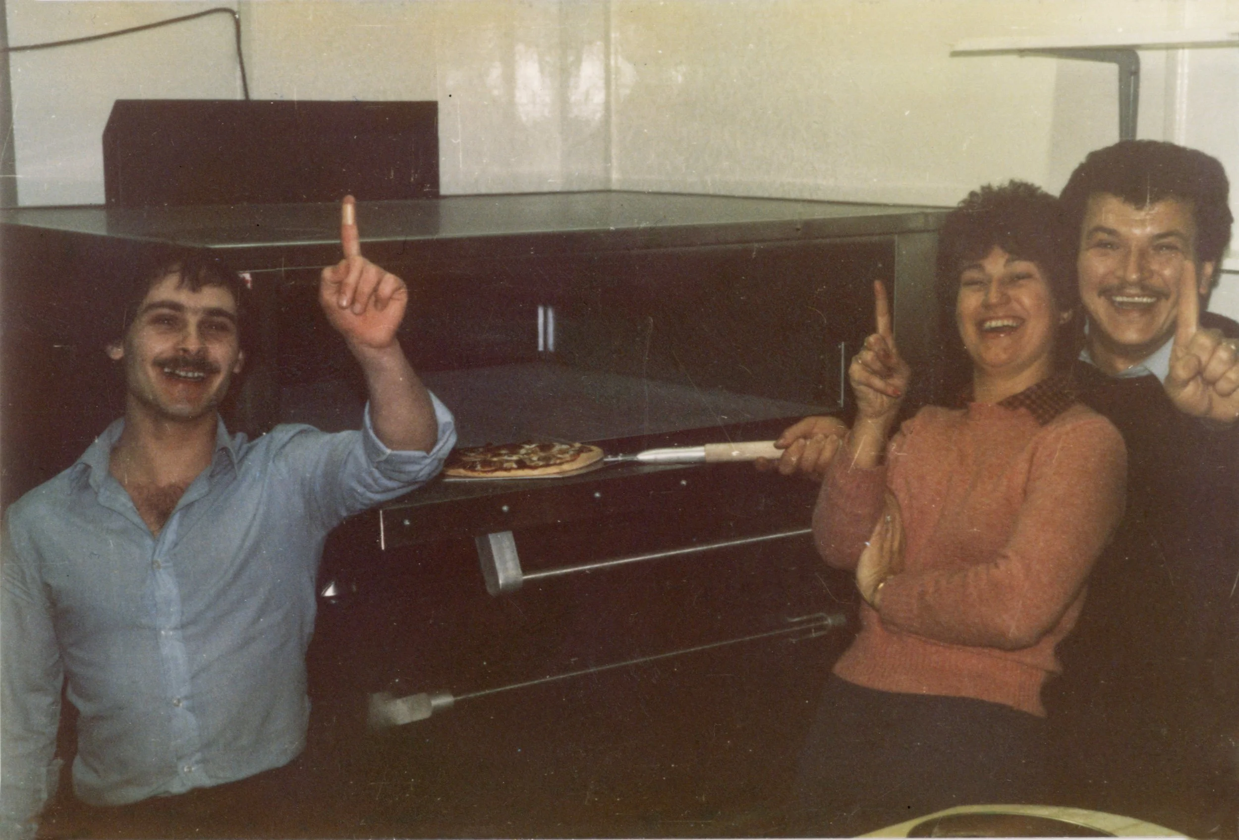 Three people are smiling and posing with their index fingers pointing upward in front of a stove with a pizza base on it. The setting appears to be a kitchen, and the photo is from an earlier era, possibly the 1980s.