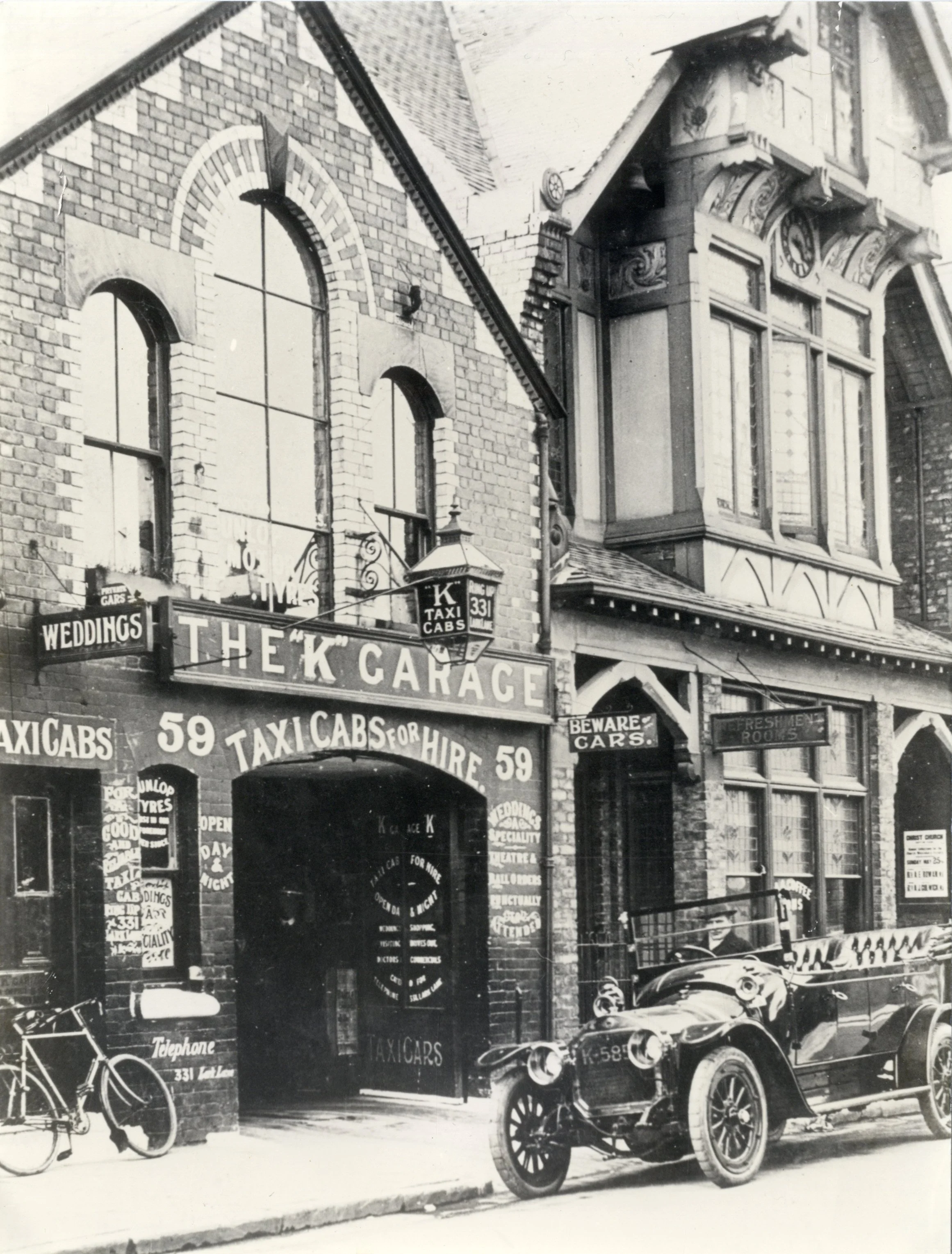 Black and white photo of a vintage corner building with brick and wood architecture, featuring signs for a taxi cab service, bicycle, and wedding services, and an old car parked in front.