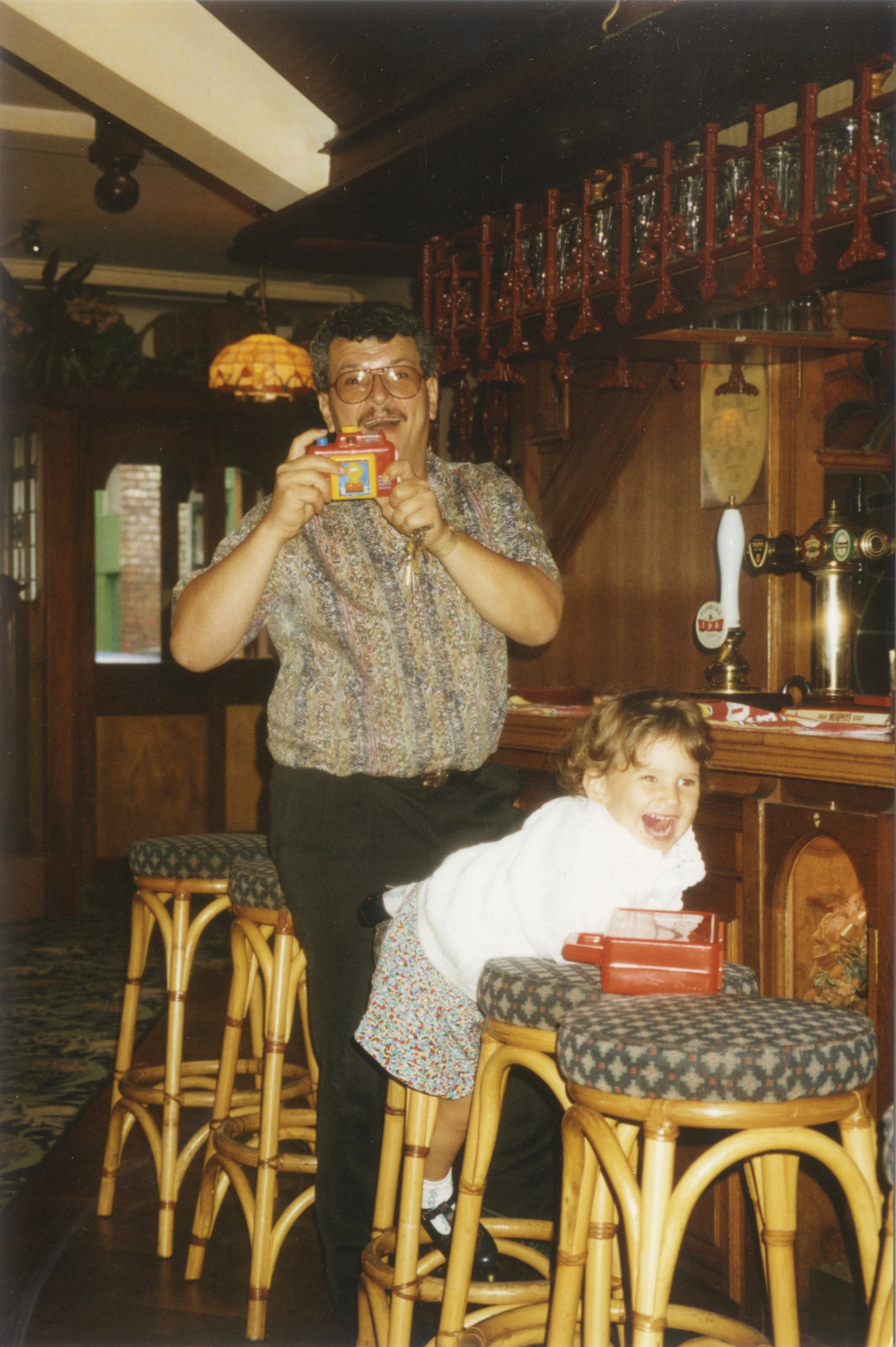 A man wearing glasses and a patterned shirt is holding a yellow camera and smiling. A young girl in a white dress with colorful patterns is laughing and leaning on a round table with a red box on it. The setting appears to be a cozy, wood-paneled restaurant or bar with glasses hanging above the bar area.