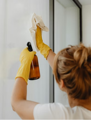 Person cleaning glass window with white cloth, wearing yellow rubber gloves, holding spray bottle.