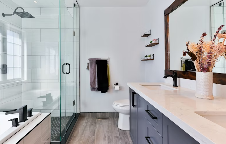 Modern bathroom with a glass shower enclosure, white tiled shower, a toilet, a blue vanity with a white countertop, a large mirror, a vase with flowers, and wall-mounted shelves with decor.