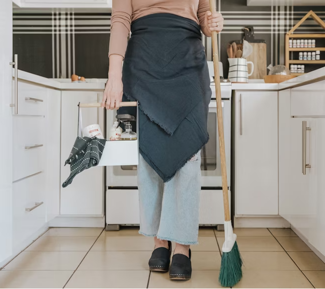Person in a kitchen holding cleaning supplies and a broom.