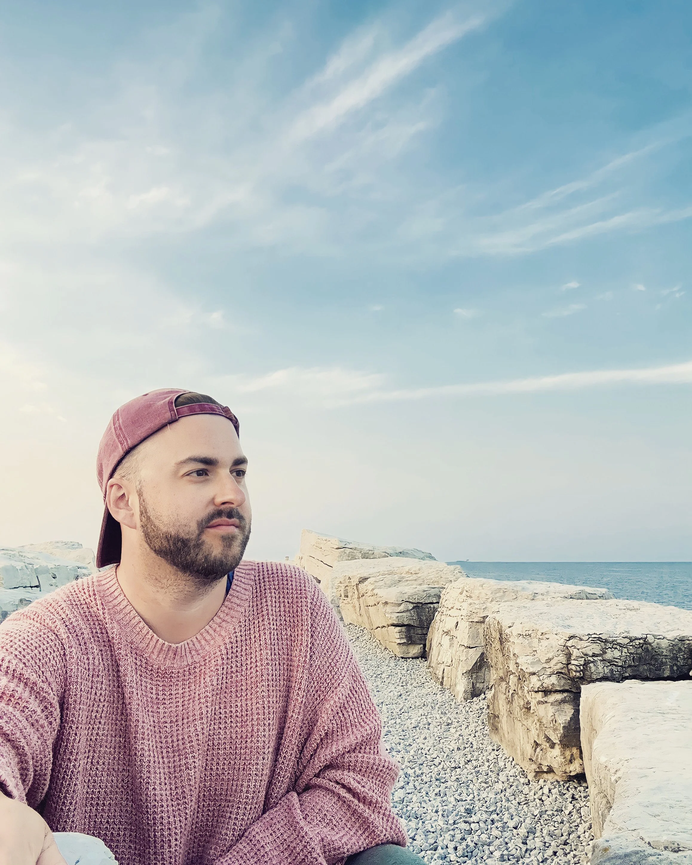 A young man with facial hair wearing a pink cap backwards and a pink sweater sitting on a rocky shoreline with large rocks and a view of the ocean under a partly cloudy sky.