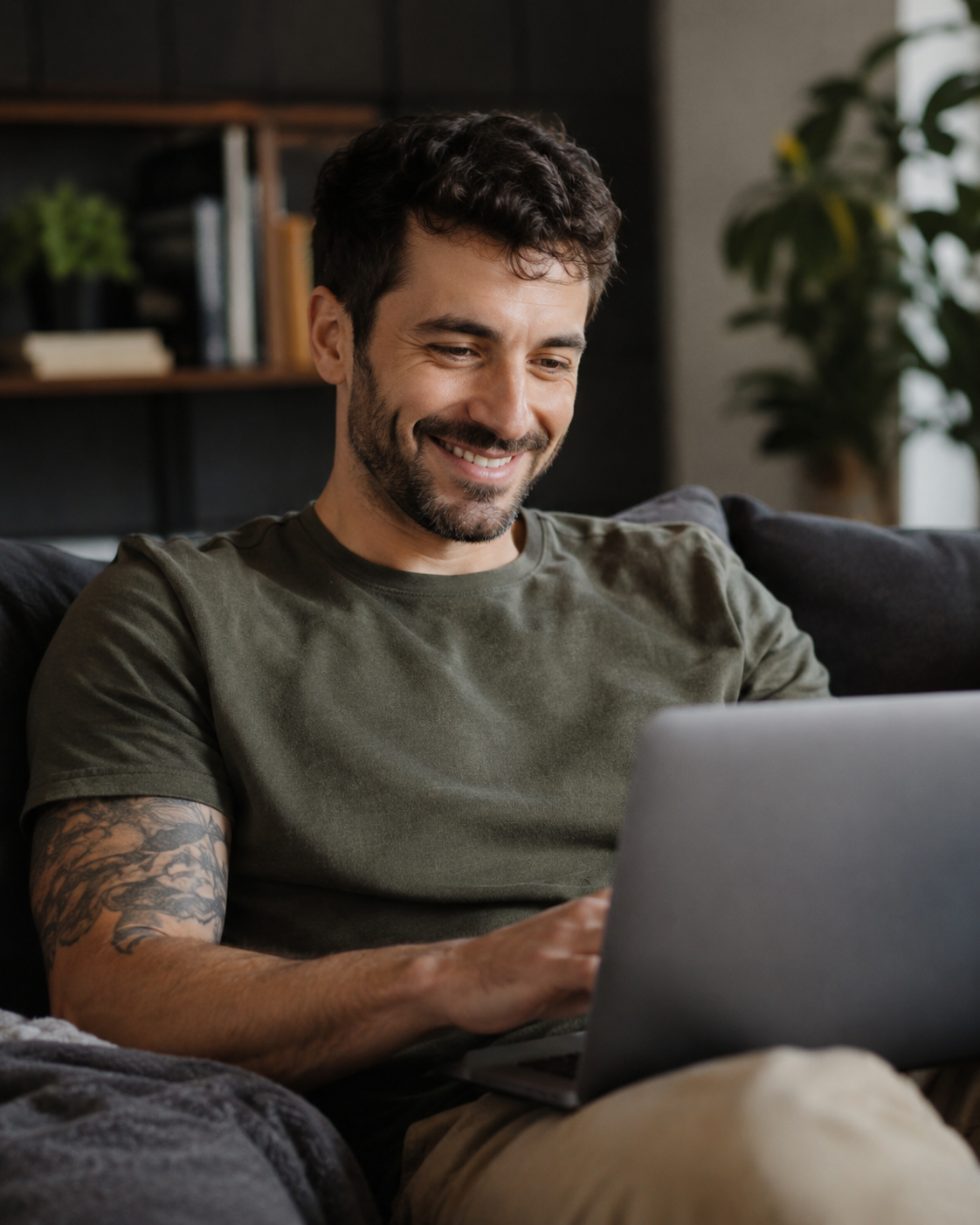 A man with dark hair, beard, and tattoos on his left arm smiling while looking at a laptop screen in a cozy living room.