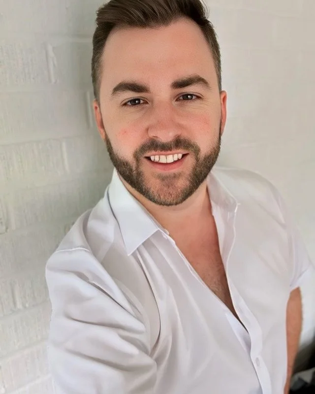 A young man with dark hair, a beard, and light skin, smiling and wearing a white button-up shirt, standing against a white brick wall.