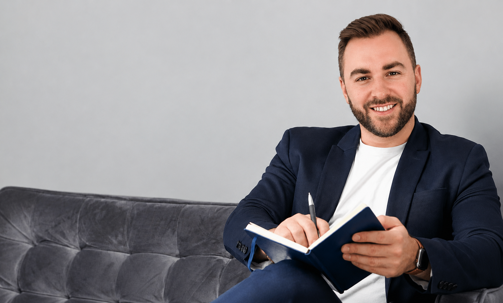 A smiling man with a beard and short brown hair, dressed in a navy blazer and white shirt, sitting on a gray couch, holding a pen and writing in a small notebook.