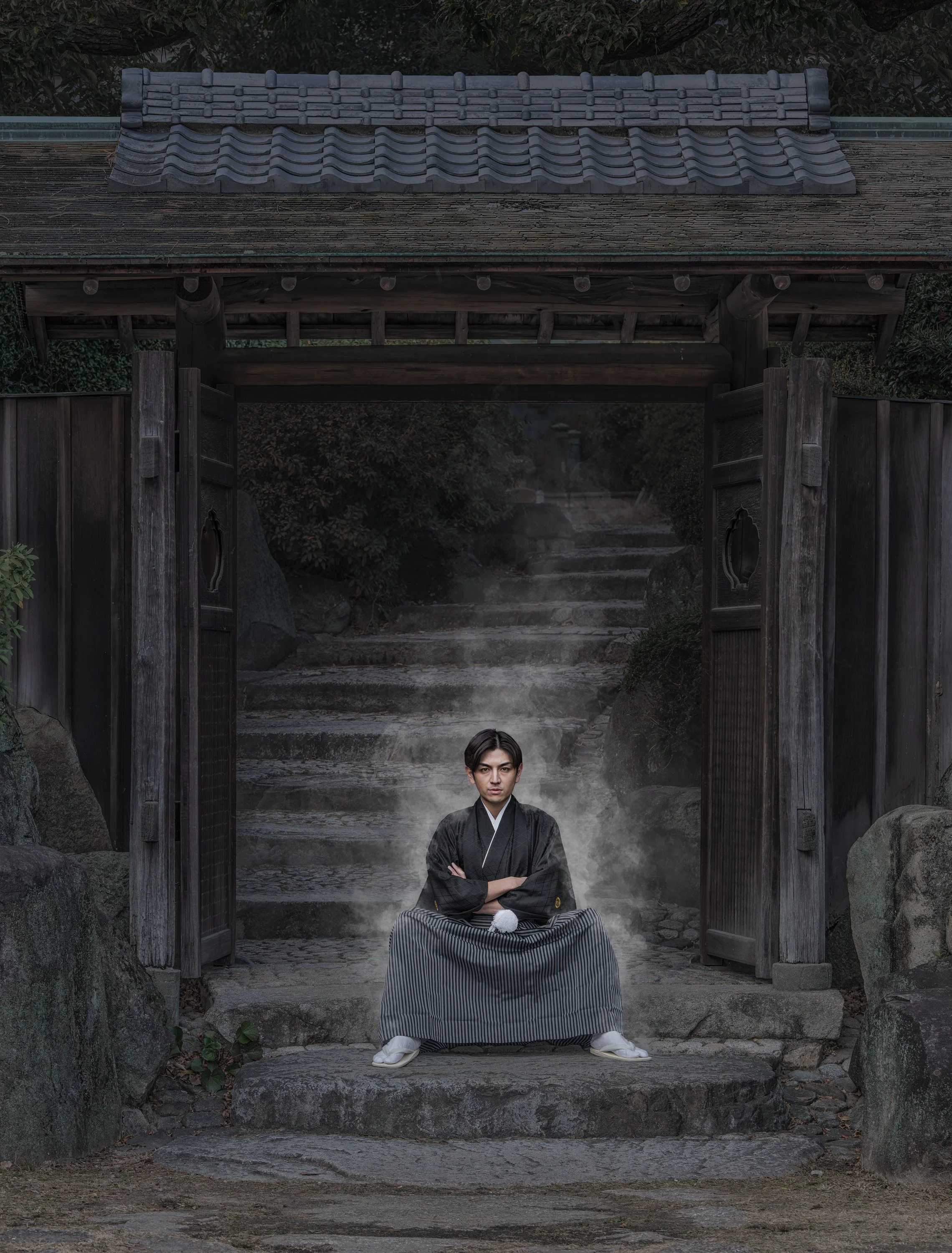 A young man dressed in traditional Japanese attire sitting cross-legged on stone steps, with steam rising around him, in front of a wooden gate and lush greenery.