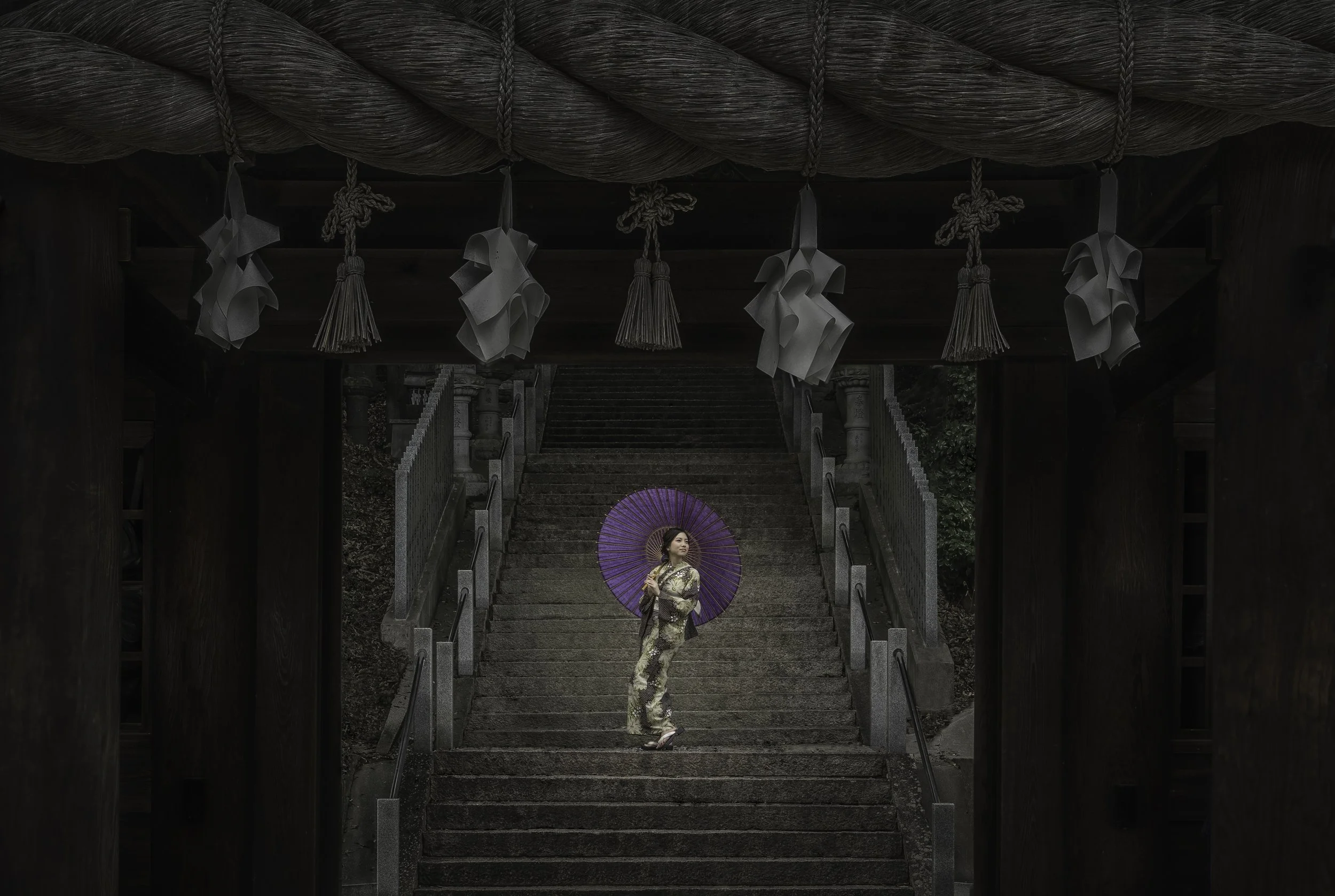A woman stands on stone steps holding a purple paper parasol, wearing a traditional kimono, in front of a Japanese shrine entrance with shimenawa rope and hanging paper shide.