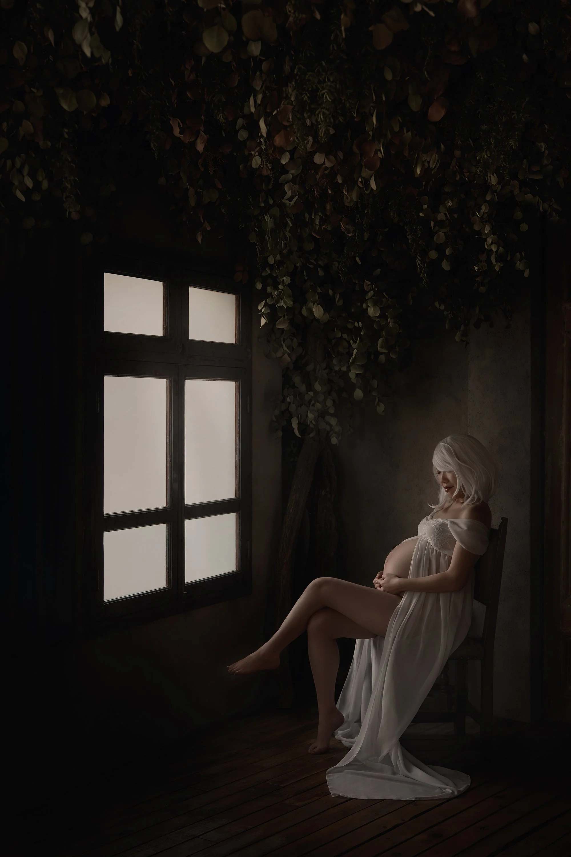 A pregnant woman with platinum blonde hair sits on a wooden chair in a dimly lit room by a window, wearing a flowing white dress.