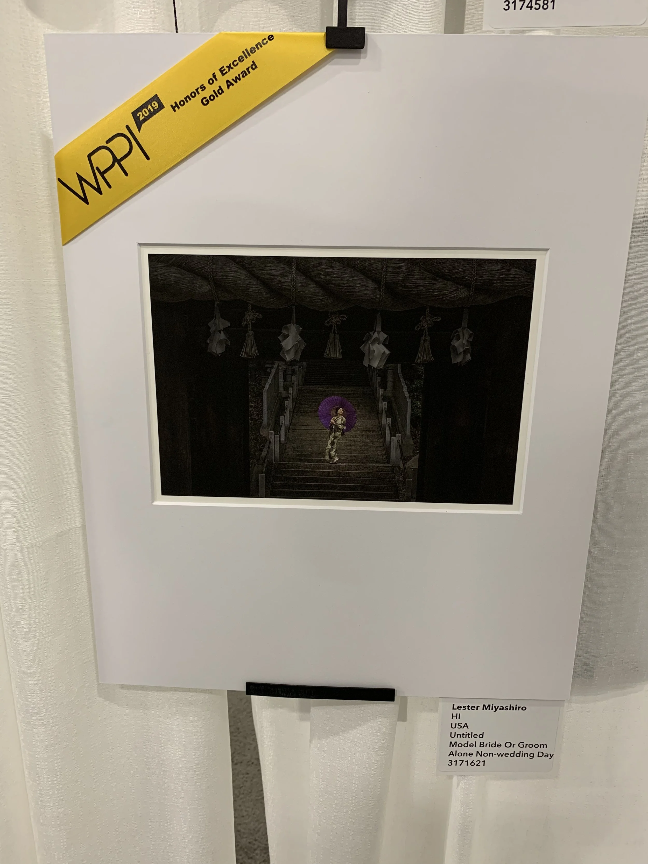 A photograph of a framed black-and-white picture of a person standing on a staircase holding a purple parasol. The staircase is decorated with hanging paper ornaments, and the background appears dark.