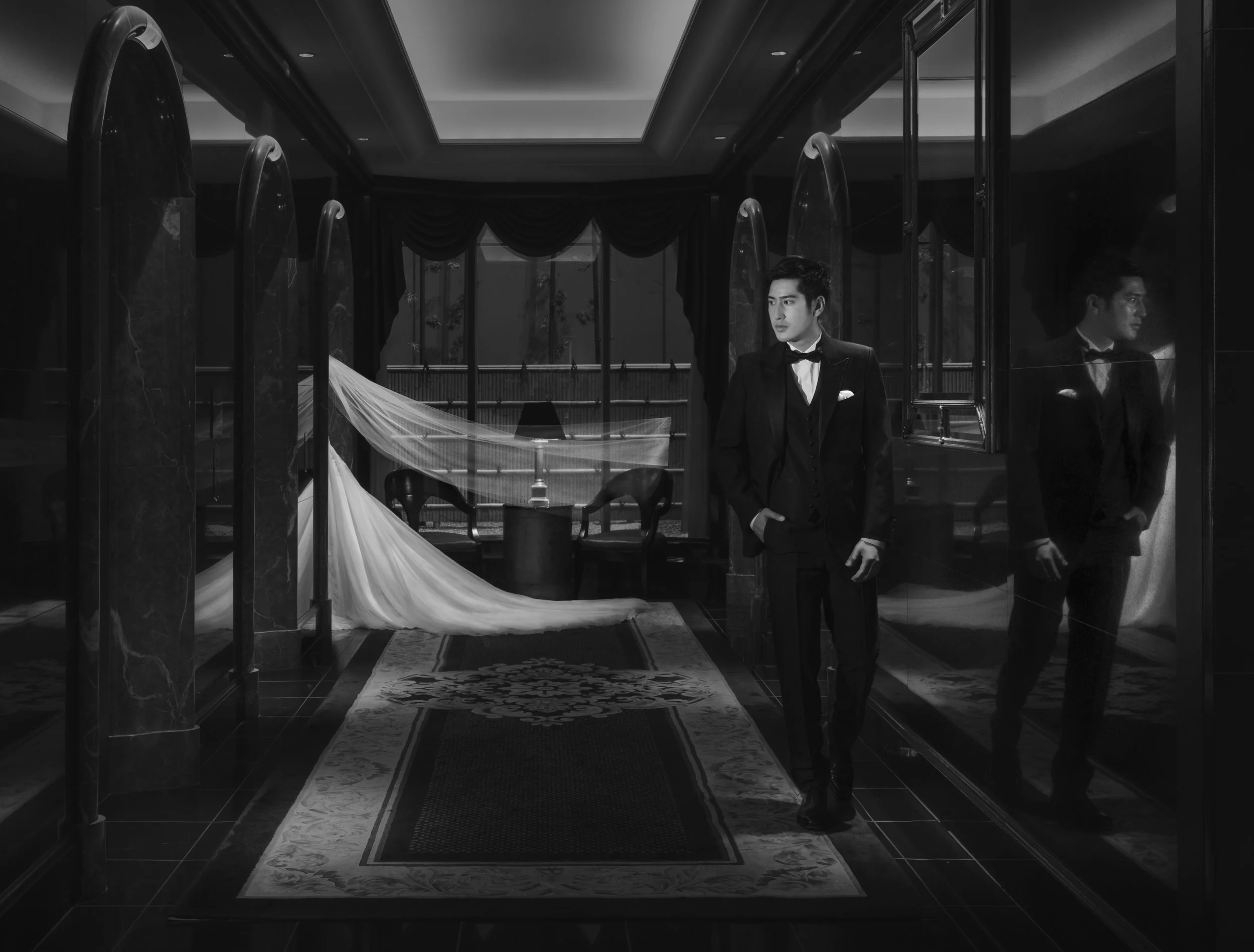 A man in a tuxedo standing in front of a glass wall, with his reflection visible, in an elegant room with dark curtains and a rug.