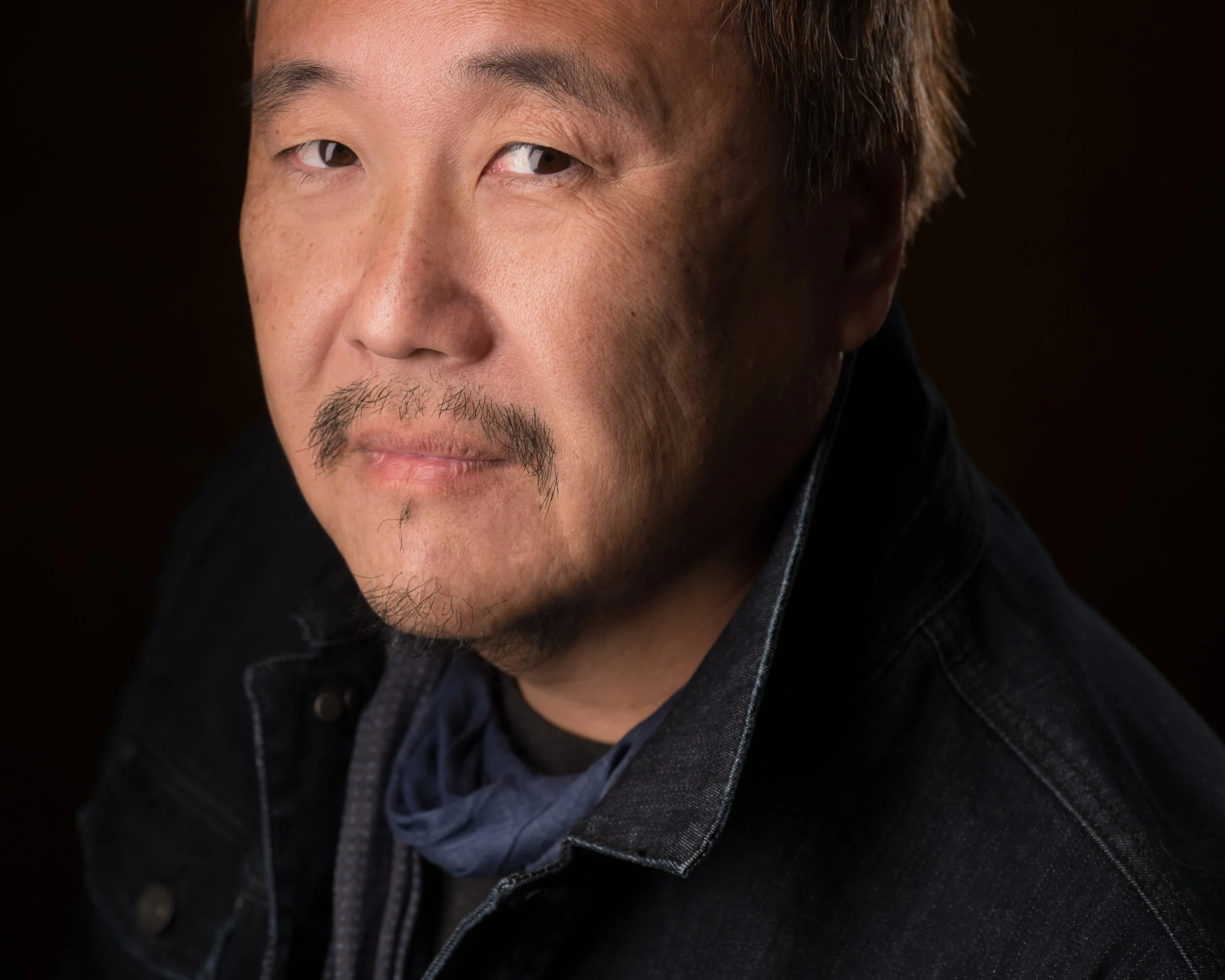 A close-up portrait of a man with dark brown hair, light skin, a mustache, and a goatee, wearing a dark denim jacket and a dark scarf, looking directly at the camera with a serious expression against a black background.