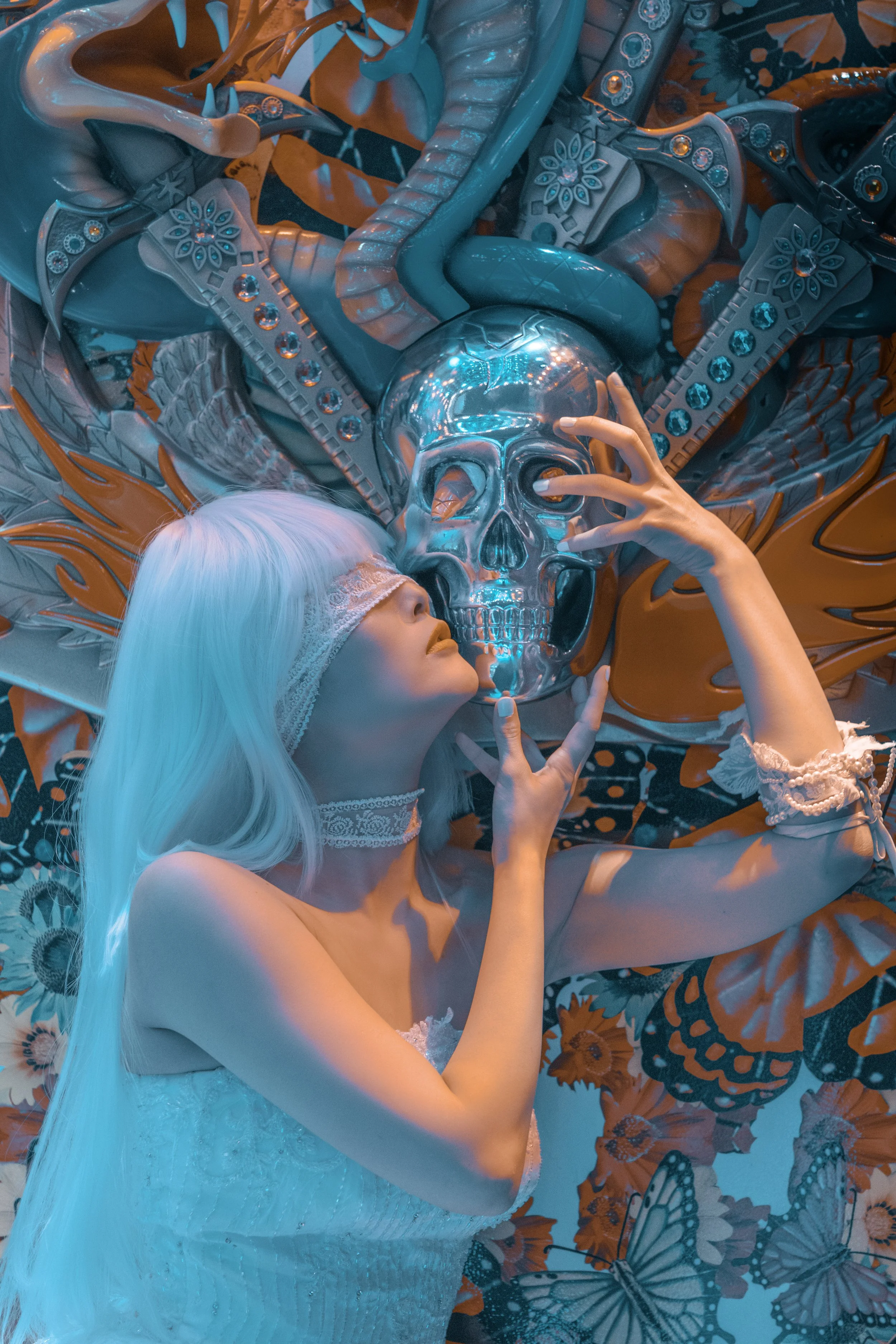 A woman with long white hair, wearing a lace blindfold and lace choker, is lying on a patterned surface. She is holding a metallic skull with one hand and touching her lips with the other. The scene is lit with blue and orange lighting, with decorative elements like butterflies and floral patterns surrounding her.