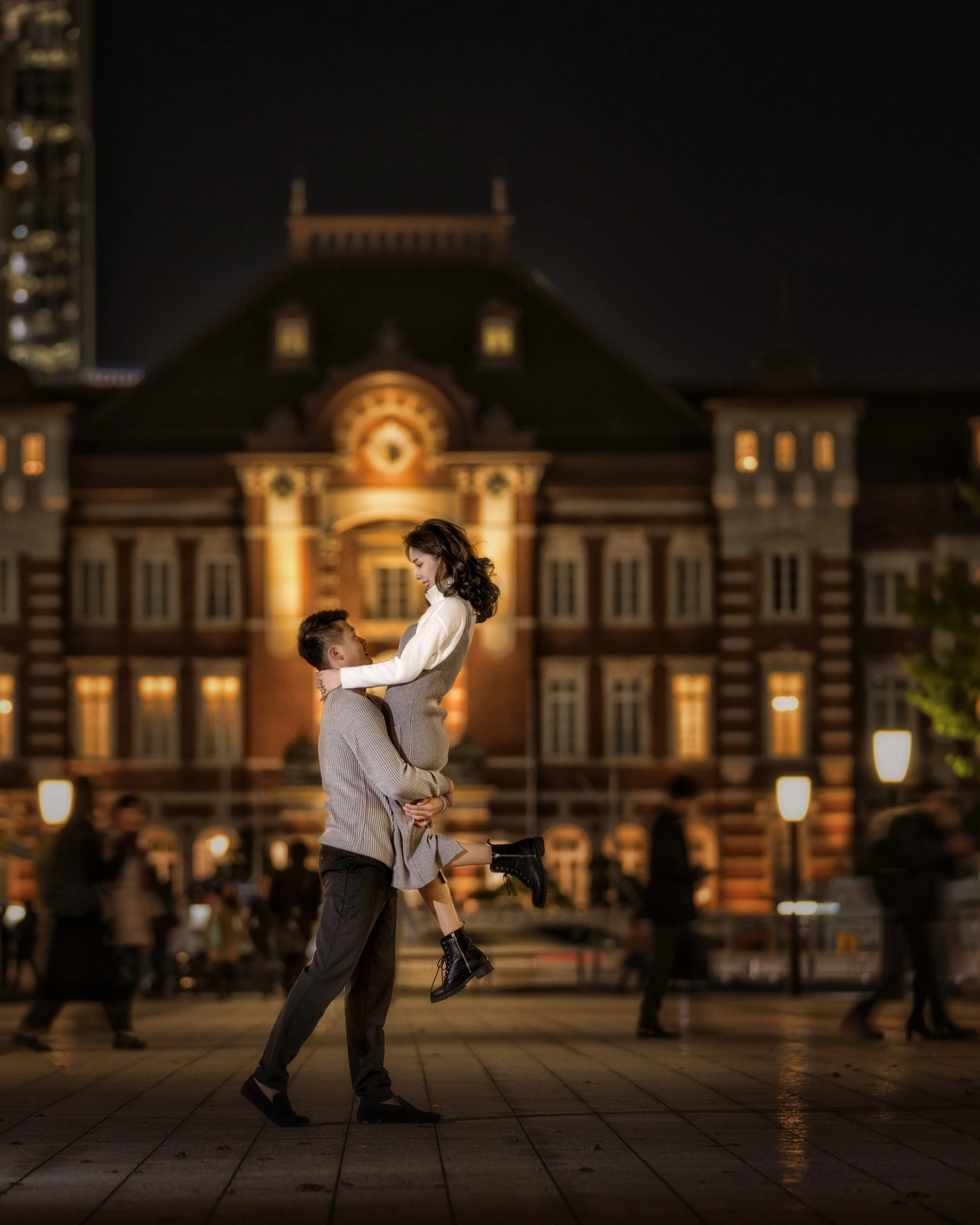 A man holding a woman in his arms at night in front of a lit historic building with a clock, with blurred people walking by in the background.