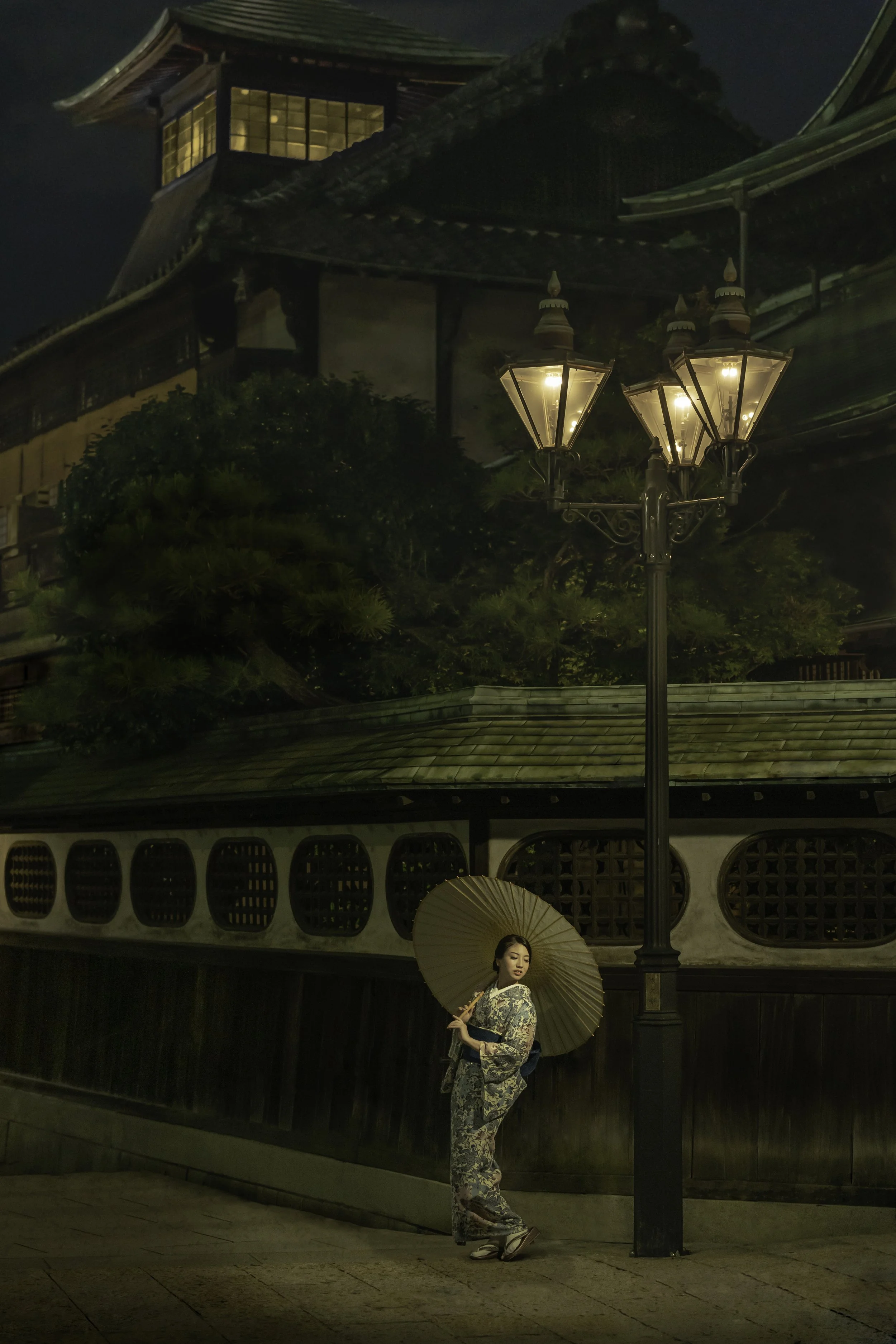 A woman in a traditional kimono holds a parasol, standing under a street lamp at night with a Japanese temple in the background.