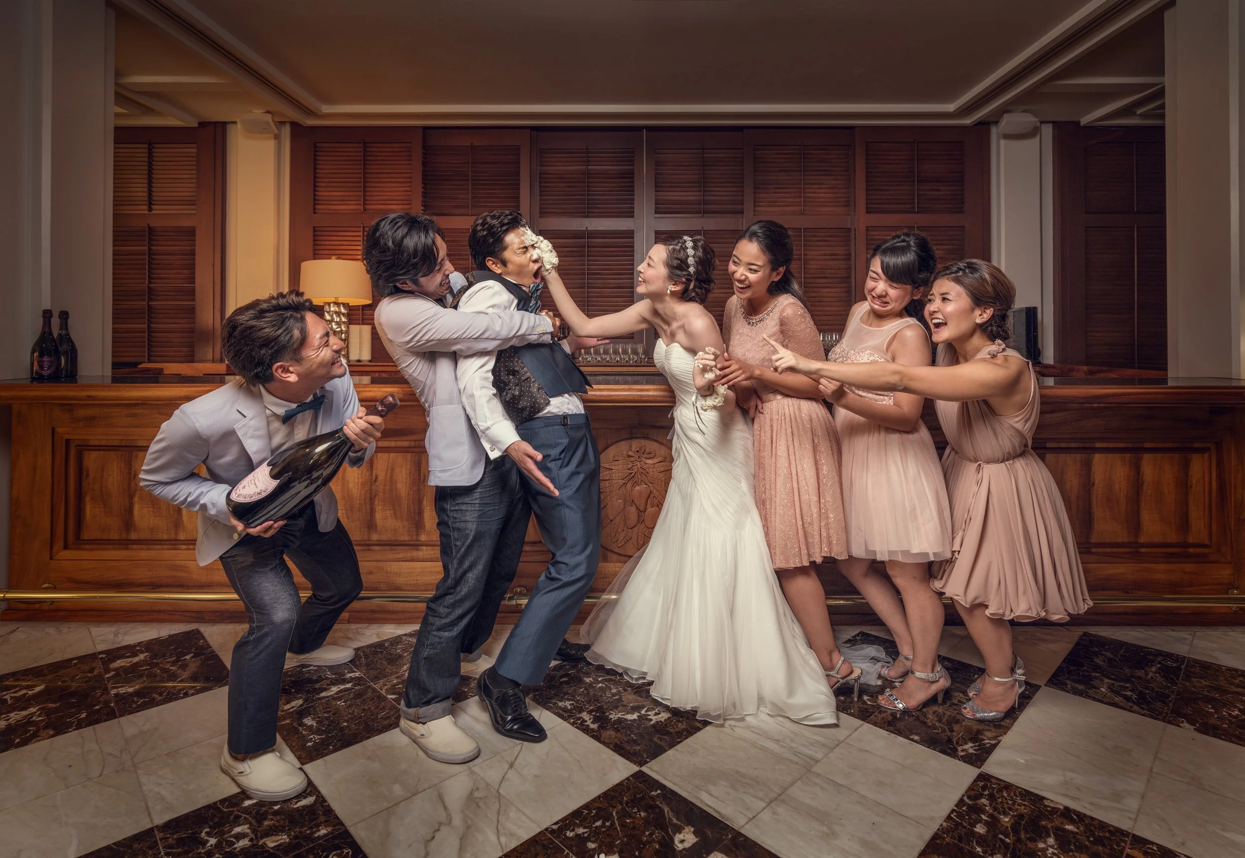 Group of people at a wedding celebration, with the bride and groom in the center, laughing and playing a playful fight as other guests join in, holding a bottle of champagne and having fun.