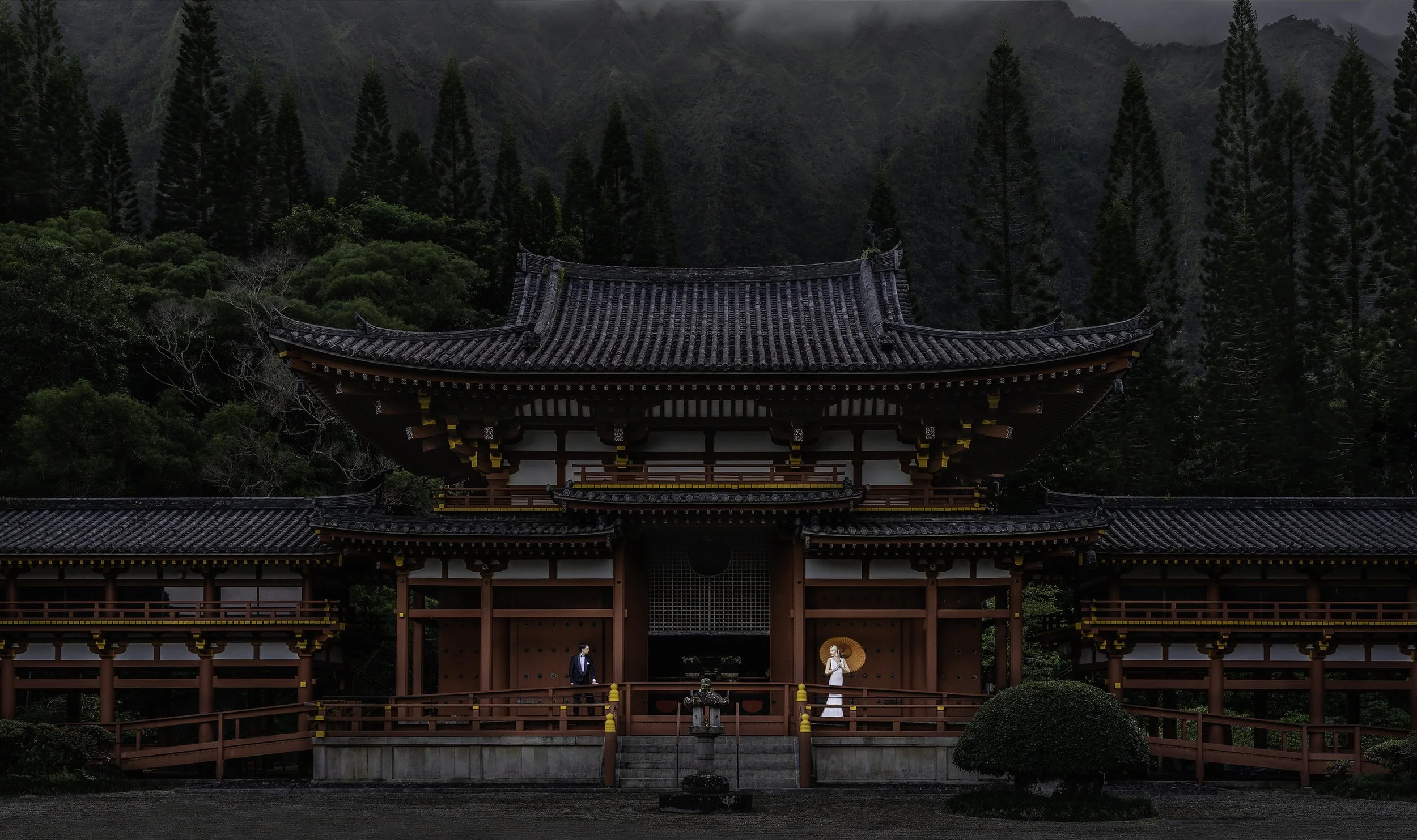 Traditional Japanese temple with red wooden structure and black tiled roof, set against a lush green mountain background. Two people, a man in a suit and a woman in a white dress holding an umbrella, stand on the temple's porch.