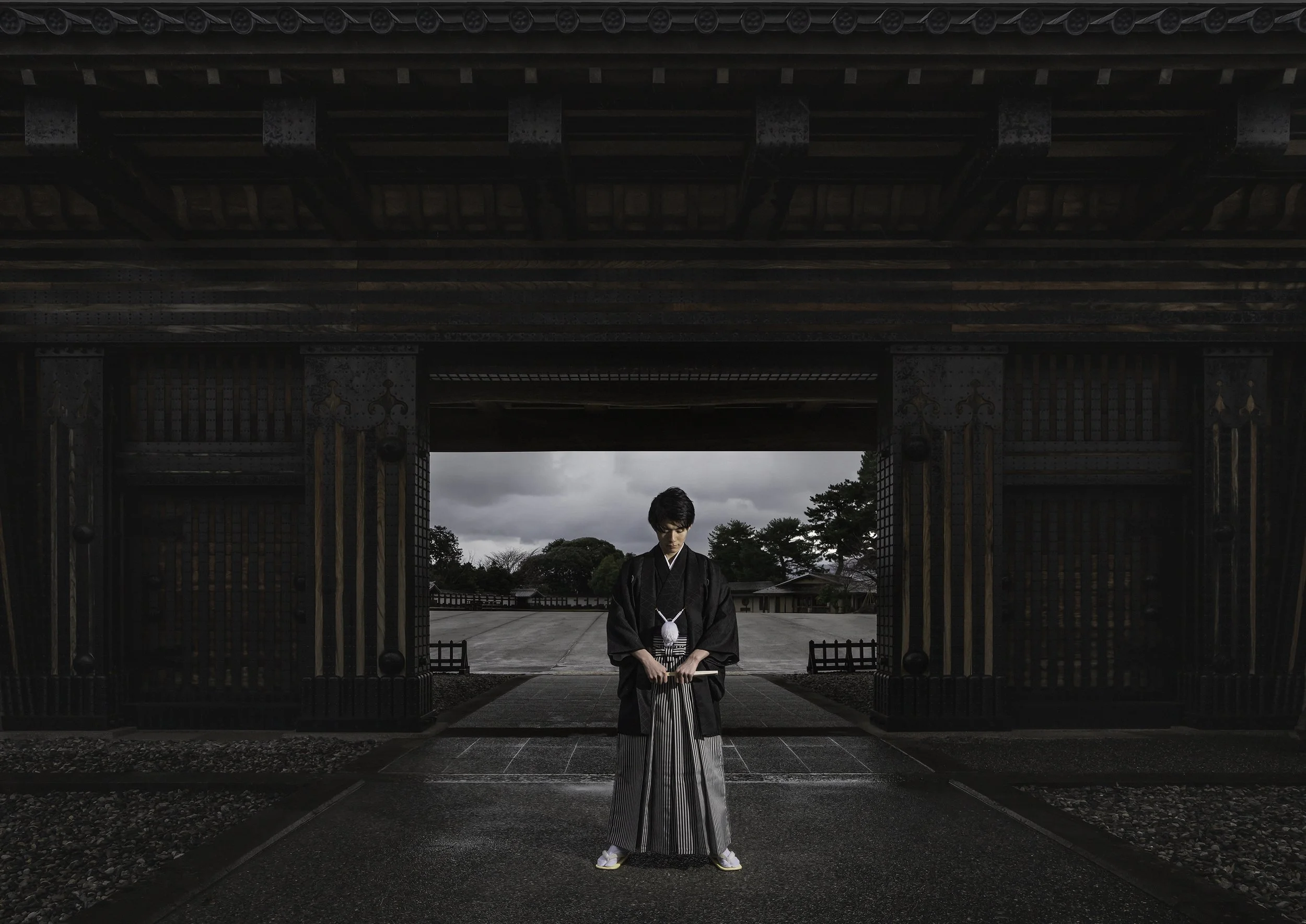A person dressed in traditional Japanese attire standing under a large ornate gate with a cloudy sky and trees in the background.