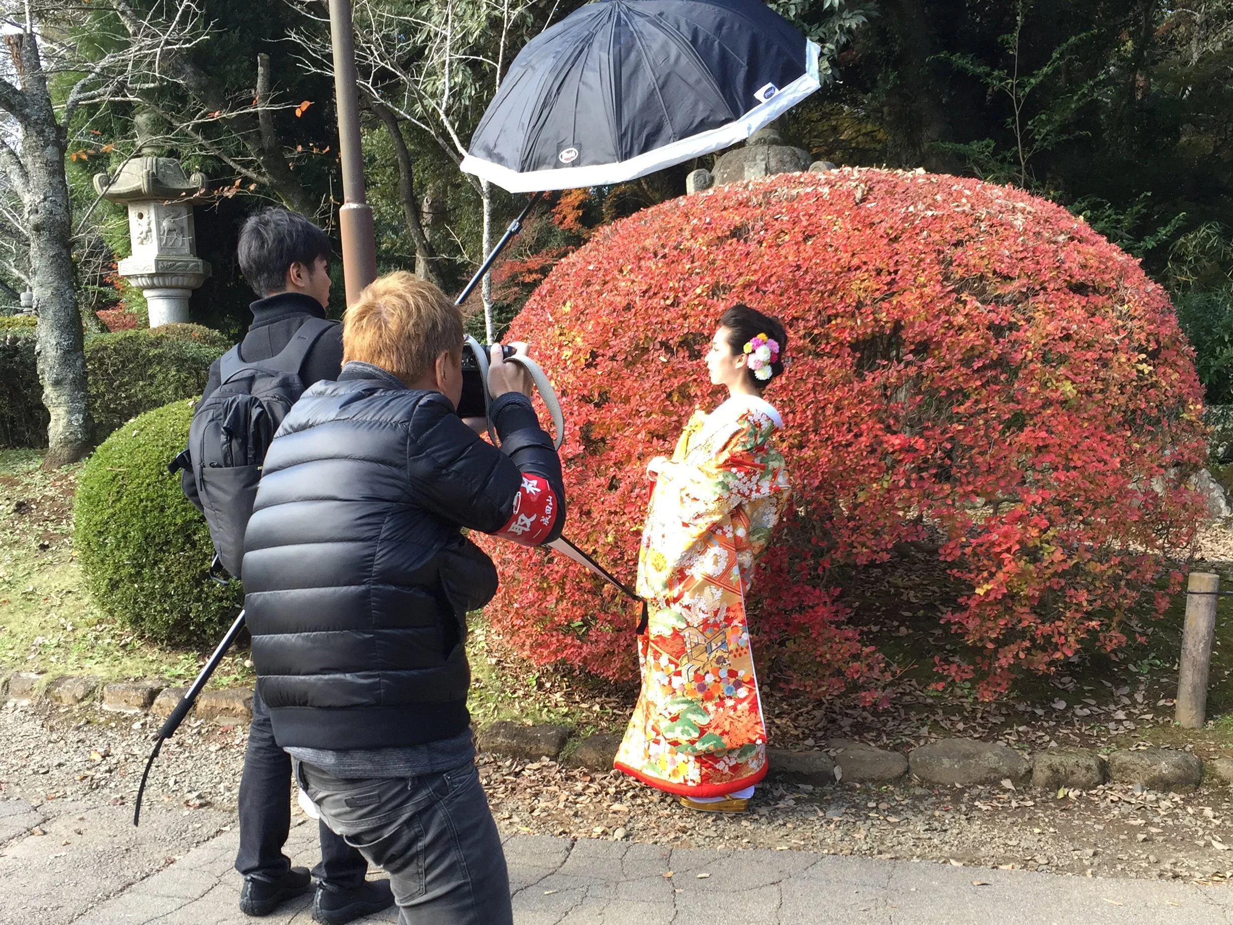 Three people photographing a woman in traditional Japanese kimono in front of a red bush during daytime.