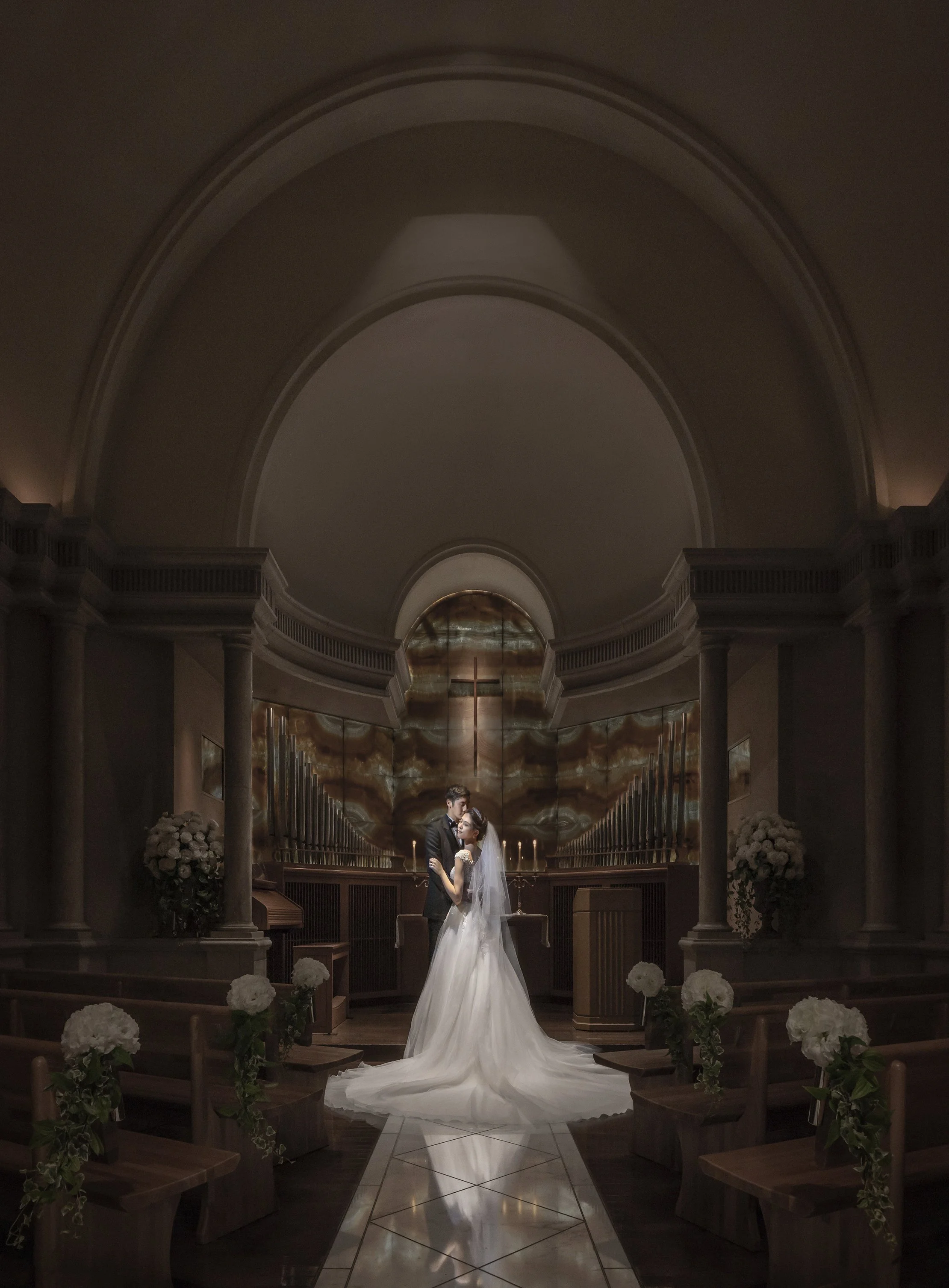 A bride and groom standing in a church, embracing each other under an altar with a cross and organ pipes, decorated with white flowers.