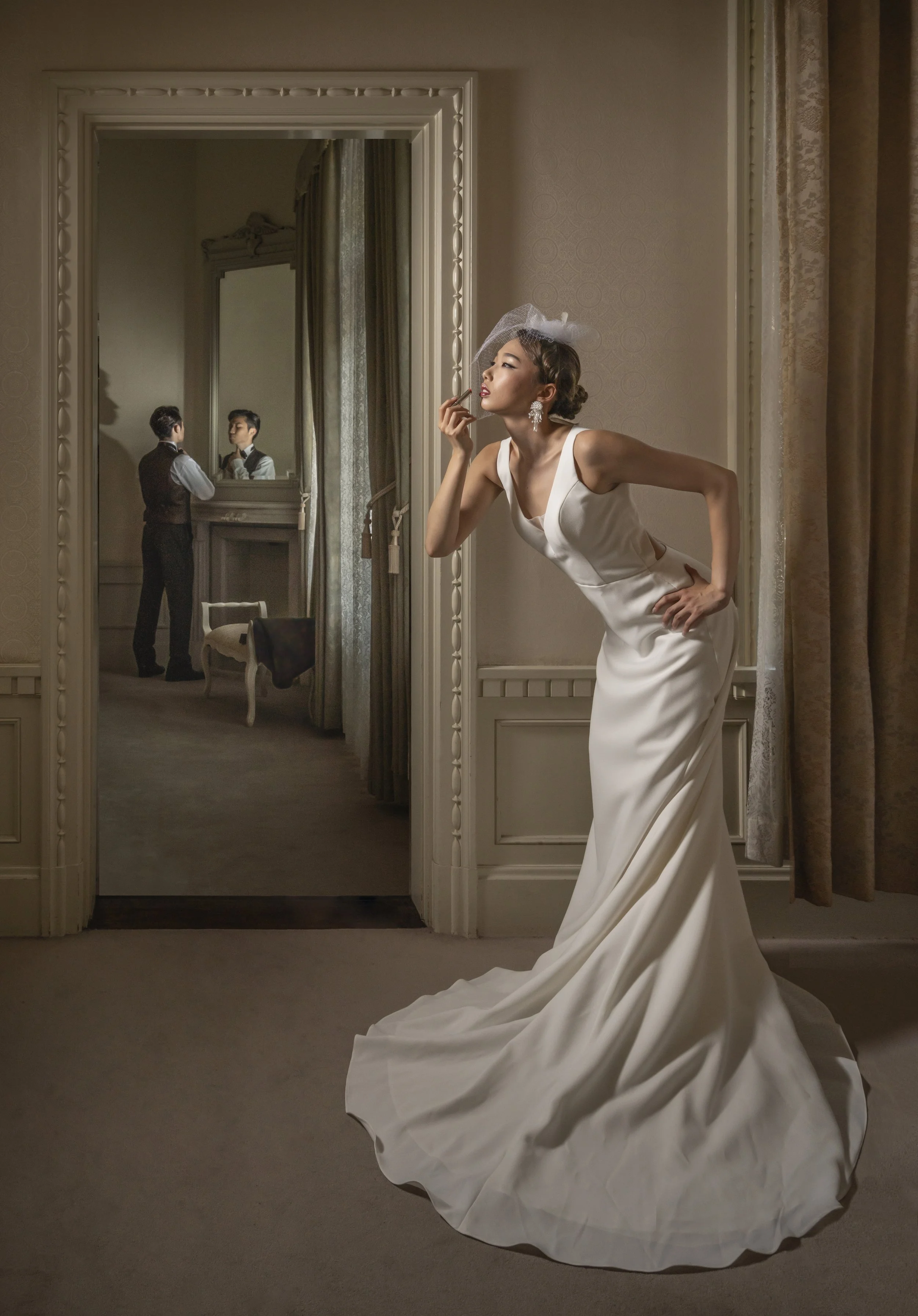 A woman in a white wedding dress and veil stands in front of a mirror, applying lipstick. In the background, a man in formal attire looks at her in the mirror, reflected in a large, ornate mirror in a vintage-style room.