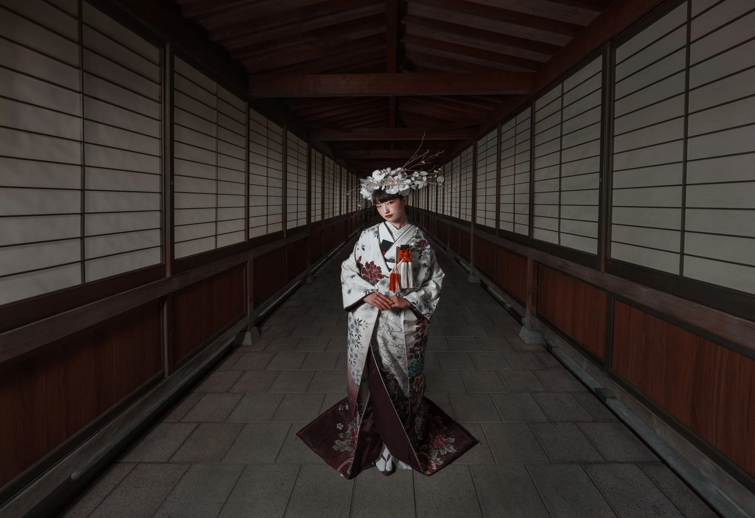 A woman in a traditional Japanese kimono standing in a covered walkway with wooden walls and shoji screens, wearing an elaborate headpiece with white flowers and branches.